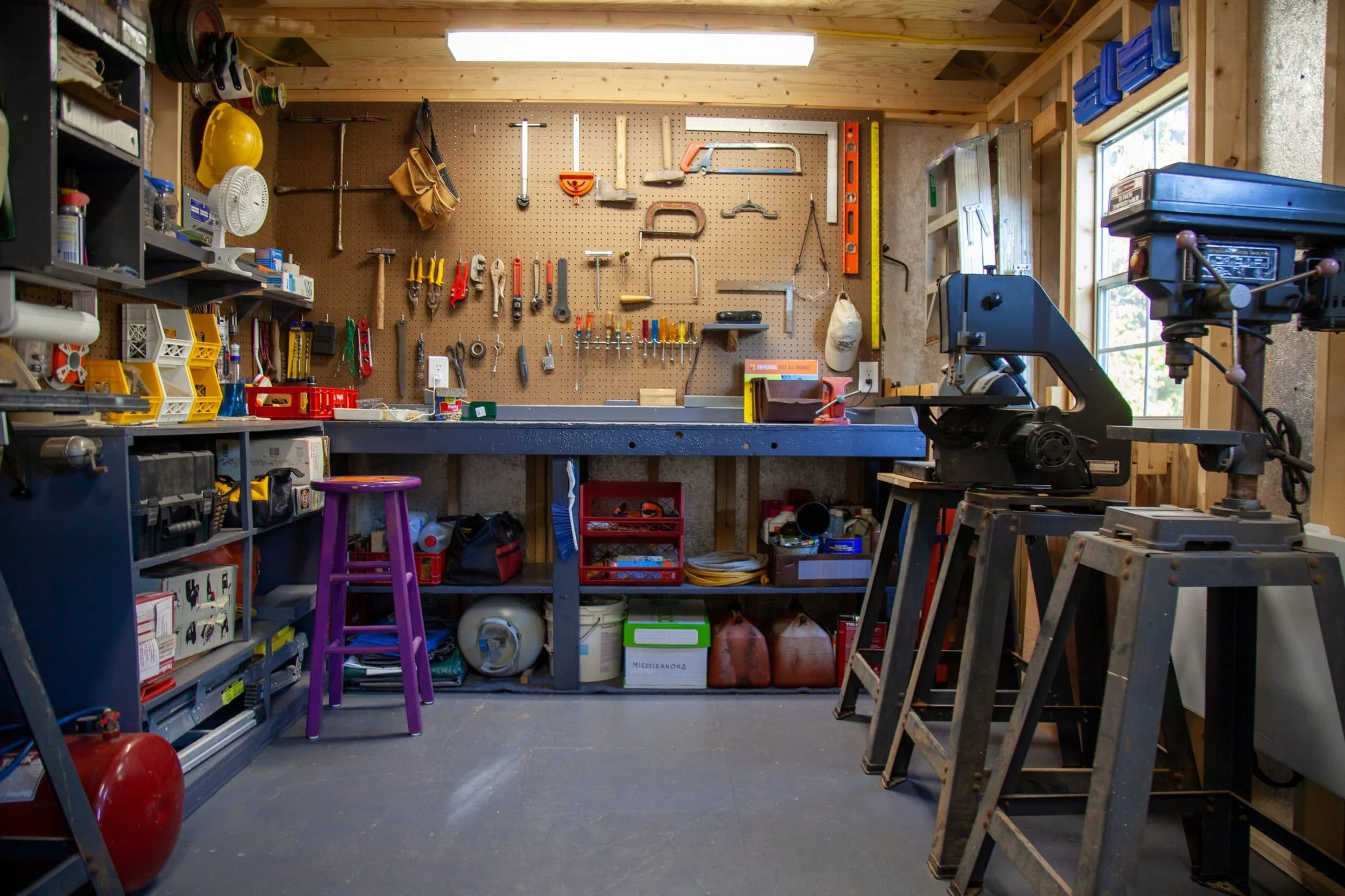 A well-organized woodworking workshop with tools hanging on the wall, workbenches, a purple stool, and various equipment and supplies stored on shelves.
