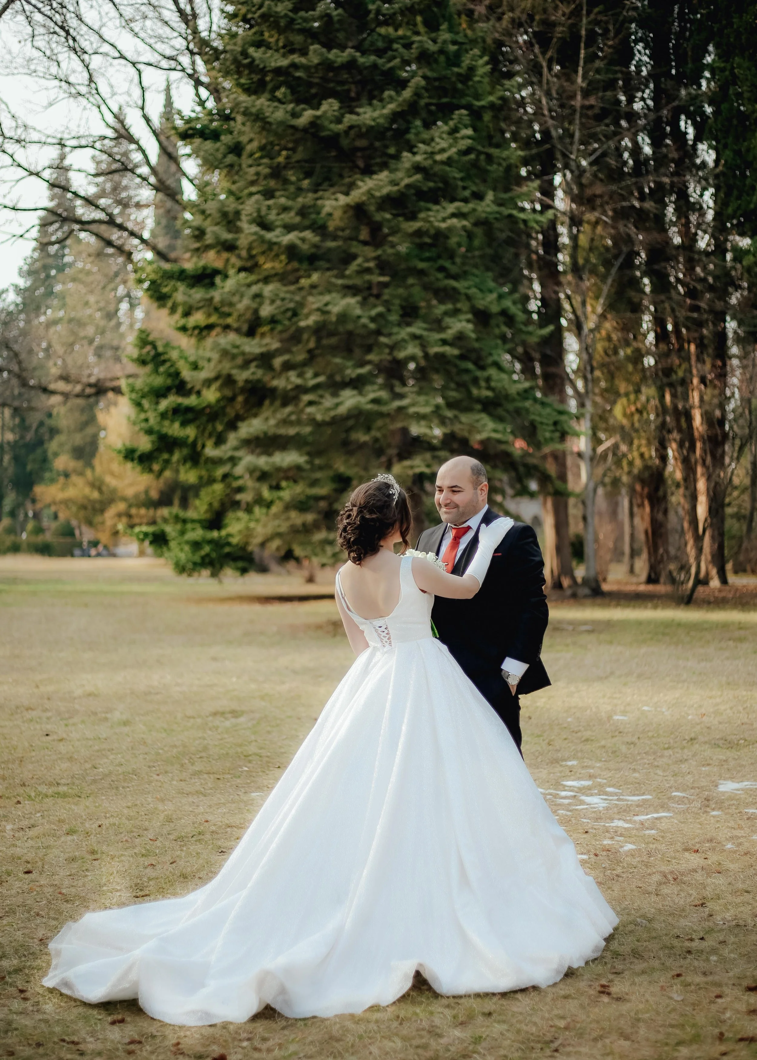 A bride and groom dancing outdoors on a grassy field with large trees in the background. The bride wears a white wedding gown, and the groom is dressed in a black tuxedo with a red tie.