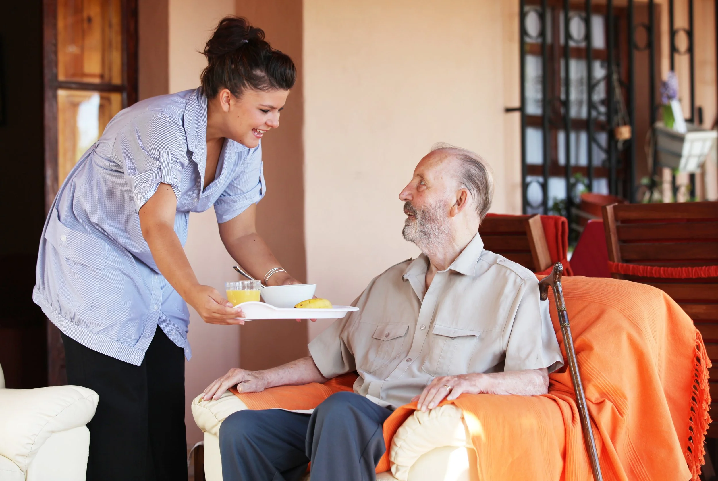 A woman is serving breakfast to an elderly man sitting on a sofa with a cane, in a cozy indoor setting.