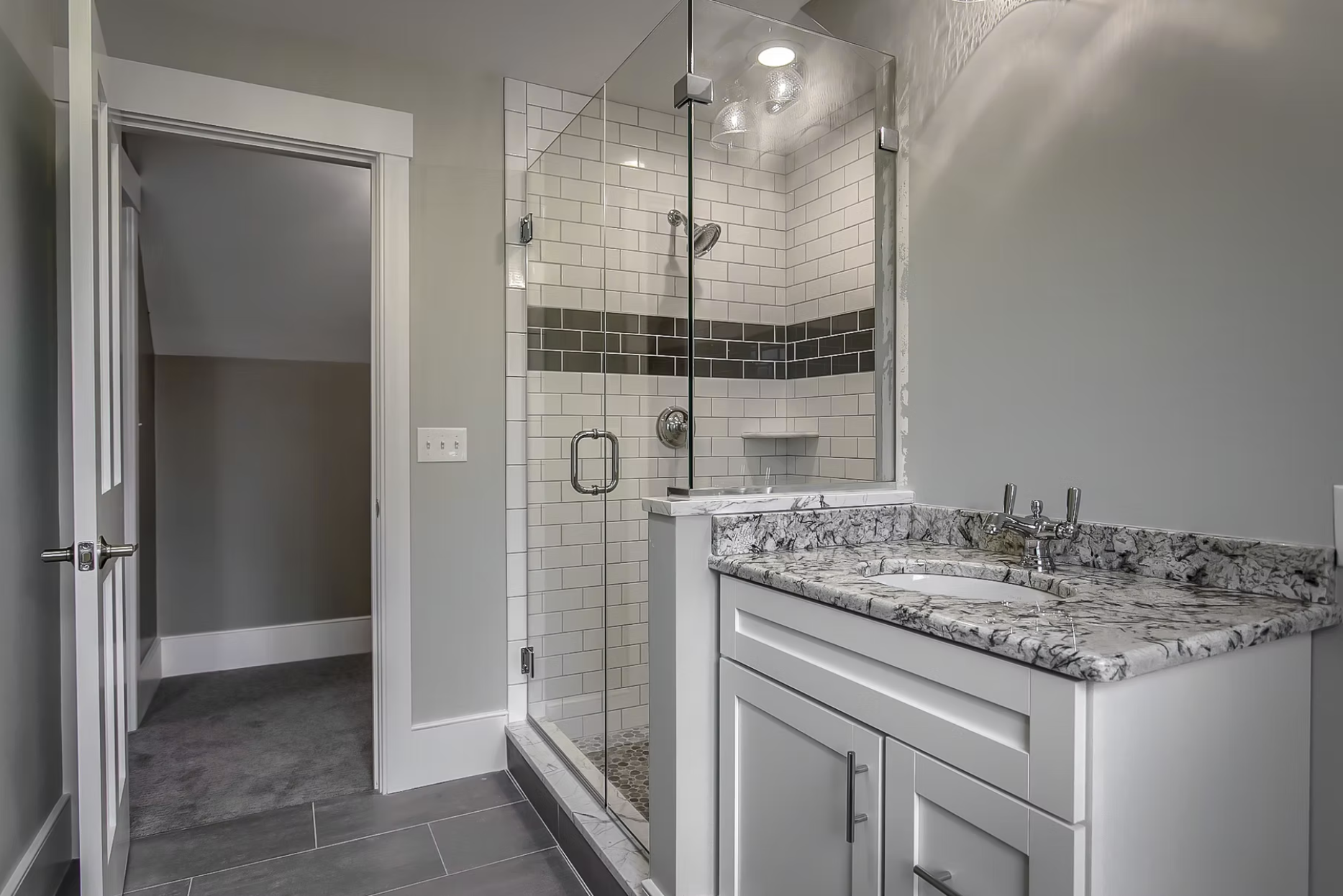Bathroom with a glass shower enclosure, white subway tile walls with a black accent stripe, marble countertop sink, gray floor tiles, and a doorway leading to a small carpeted room.