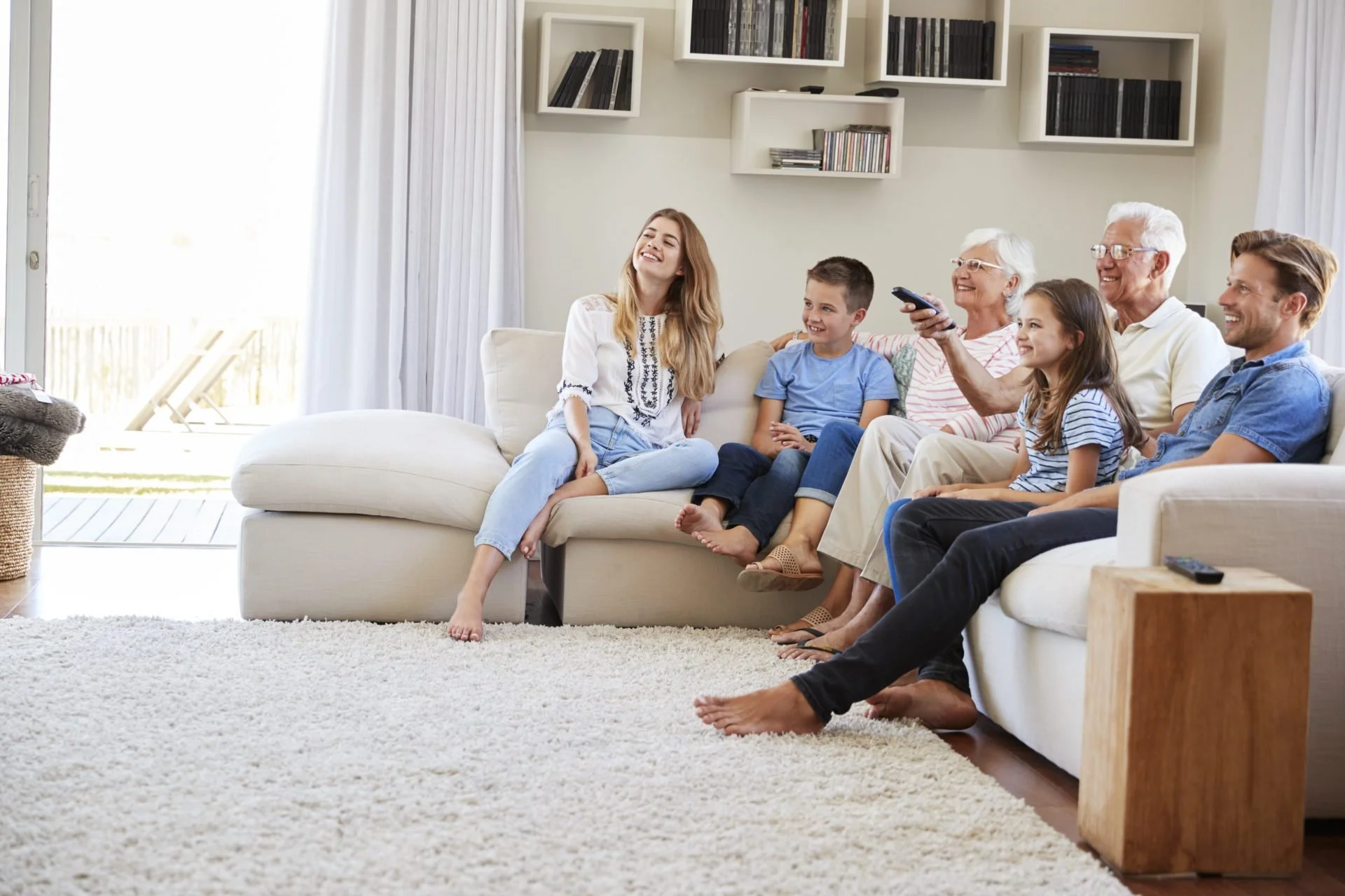 A multigenerational family sitting on a white sofa in a living room, smiling and enjoying each other's company, with sunlight coming in through large windows.