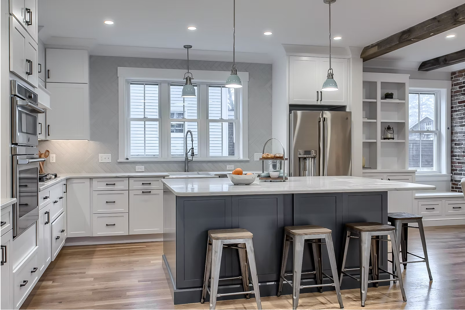 Modern kitchen with white cabinetry, a dark gray island with four stools, stainless steel refrigerator, double oven, and windows with blinds, wooden floors, and exposed wooden beams.