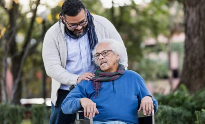 A man with glasses and a beard leaning over an elderly woman in a wheelchair, outdoors in a park.