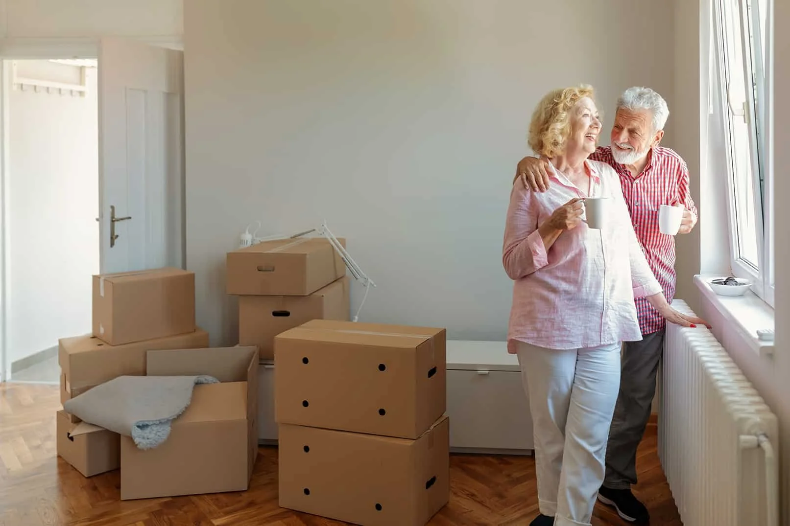 An elderly couple standing by a window, drinking coffee and smiling at each other in a room with moving boxes and packing items.