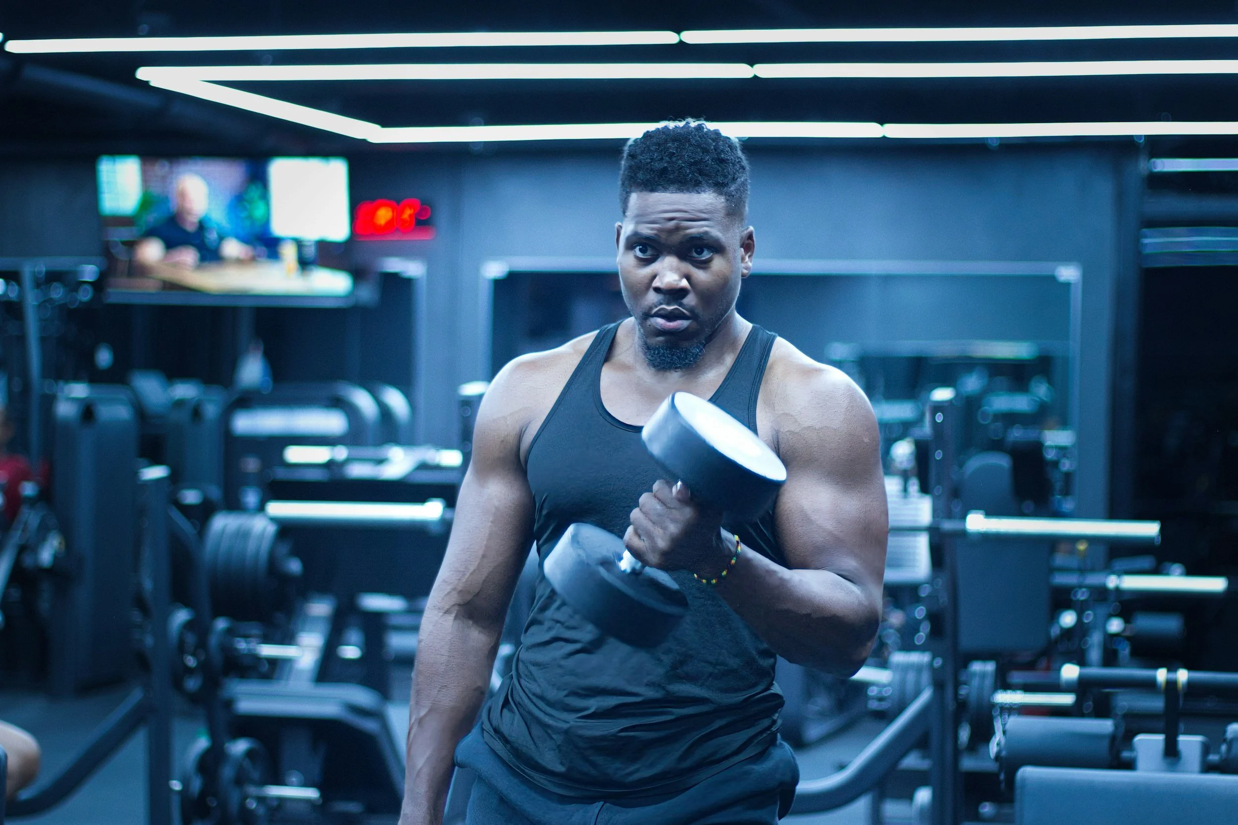 A muscular man in a black tank top lifting a dumbbell in a gym with dark walls and bright LED lighting.