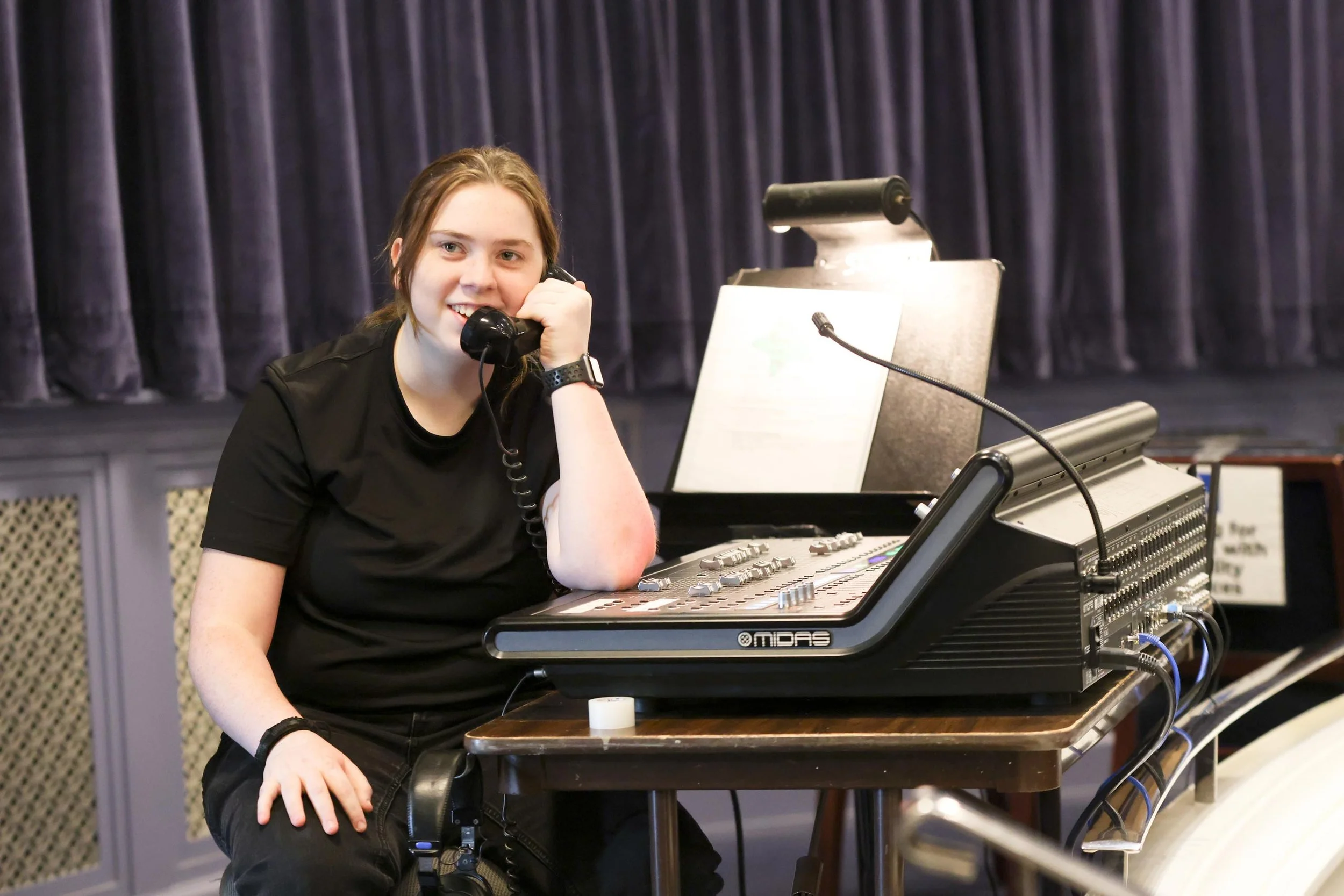 A woman with red hair sitting in front of a sound mixing console, holding a telephone receiver to her ear, with a dark curtain in the background.