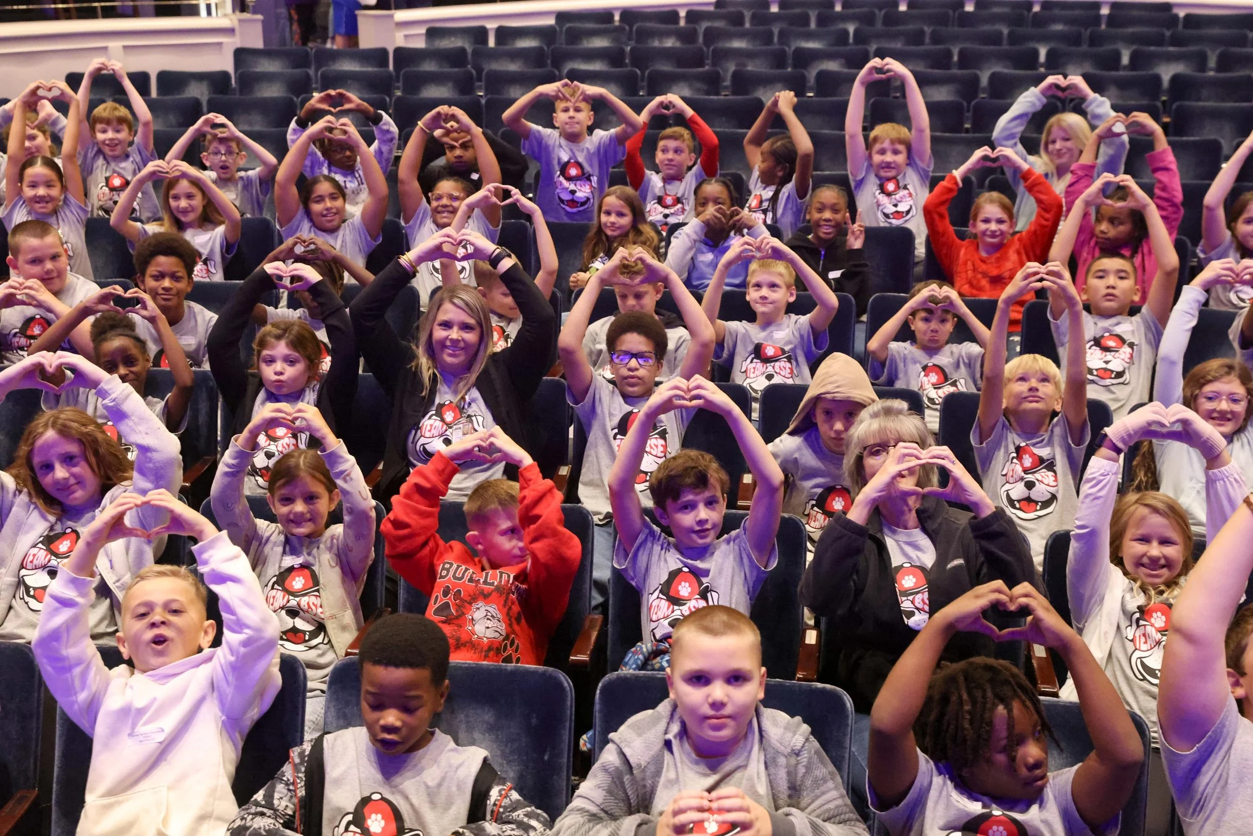 A large group of children sitting in theater seats, making heart shapes with their hands. They are wearing matching shirts with a paw print and cartoon character, indicating a school or group event.