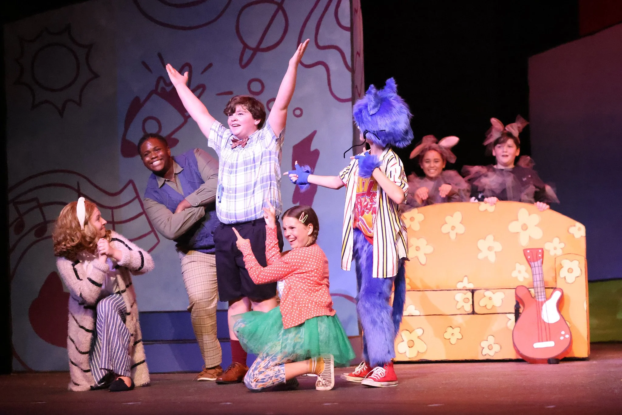 Children and performers on stage during a children's theater show, with colorful costumes, set decorations of flowers, guitar, and a license plate, engaging in animated expressions and poses.
