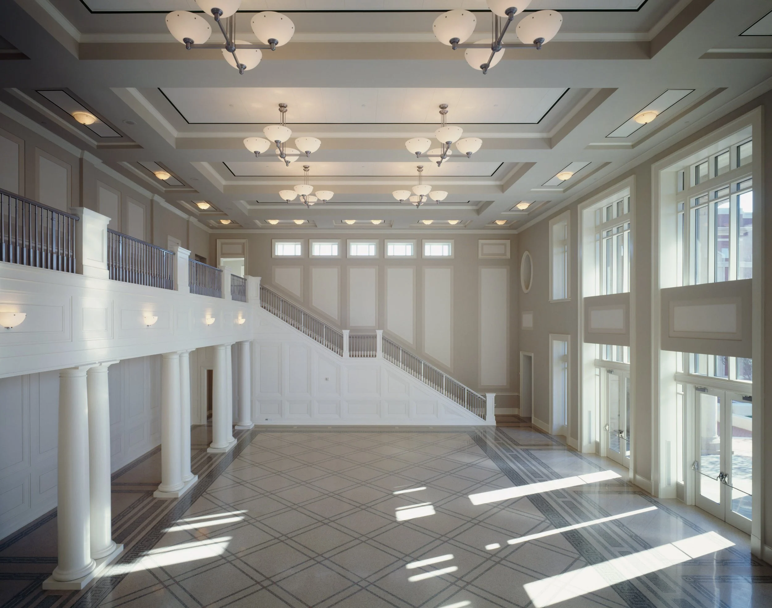 A large, spacious interior of a building with high ceilings and multiple chandeliers. Rows of windows allow natural light to fill the space, which features white walls, decorative moldings, and a tiled floor with intricate patterns. A staircase with 