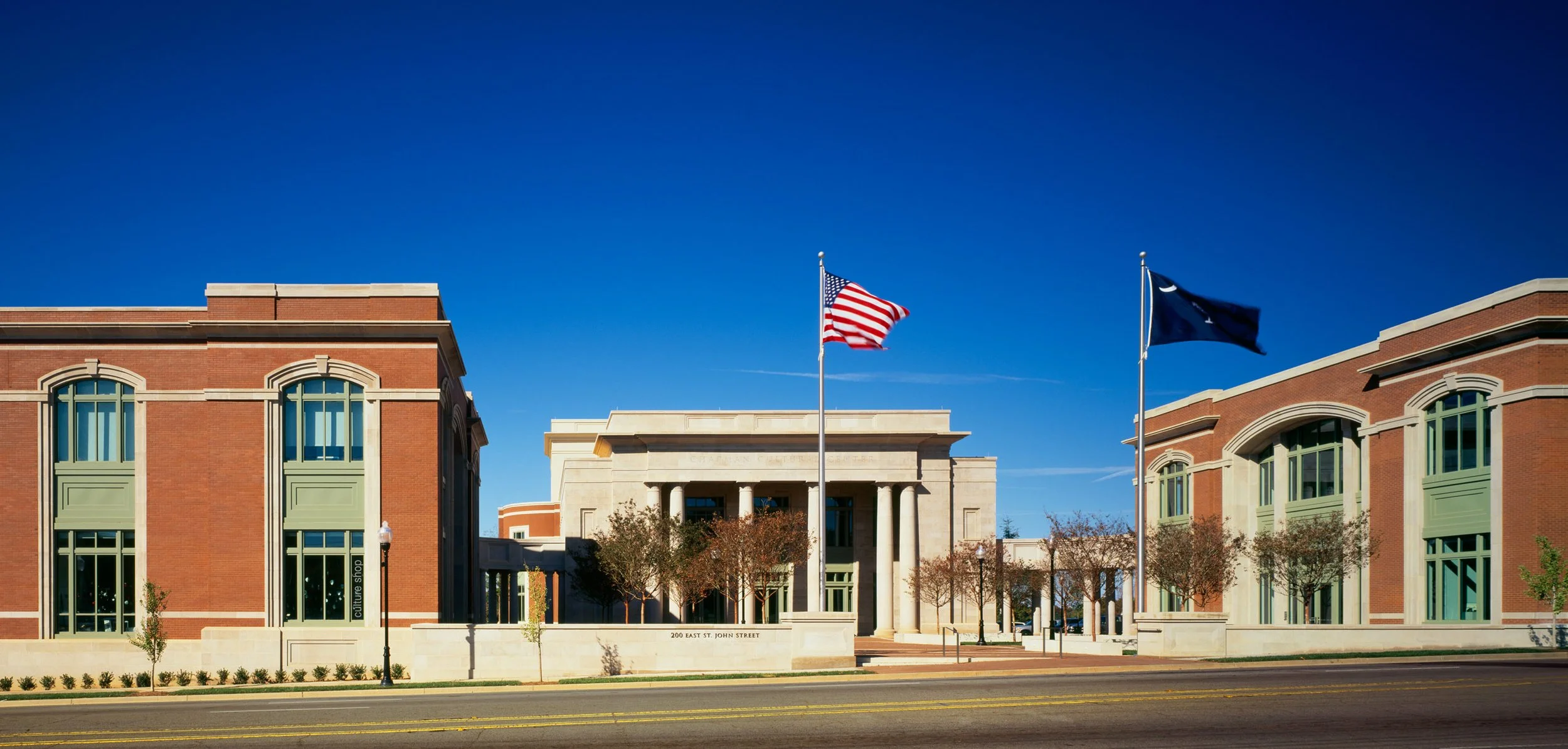 A building with red brick walls, green window trims, and a white classical portico with columns. Two flags, the American flag and a dark blue flag, are flying on flagpoles in front of the building. The street in front has a double yellow line, and sm