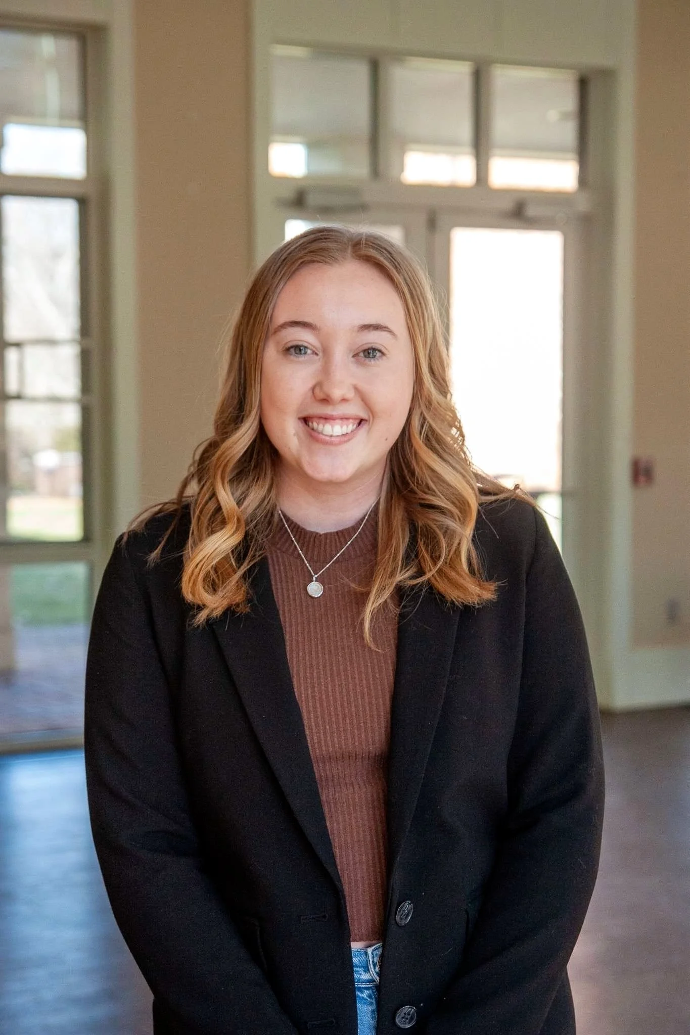 Portrait of a young woman with wavy blonde hair, wearing a black blazer and a pendant necklace, smiling, indoors with large windows in the background.