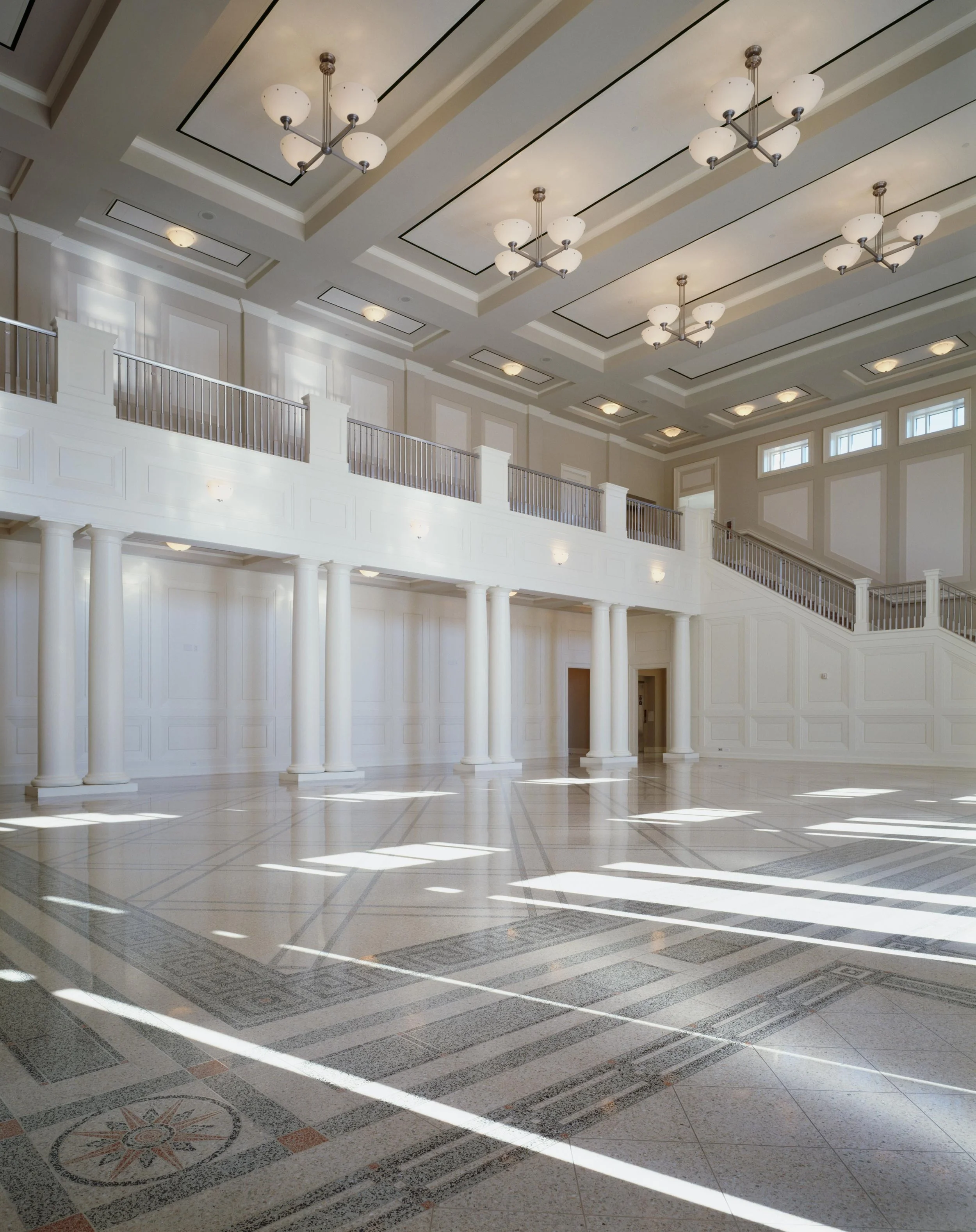 Spacious, elegant interior with a high ceiling featuring multiple modern chandeliers, white walls with decorative panels, large windows letting in natural light, granite flooring with a detailed geometric pattern, and a balcony with white columns.