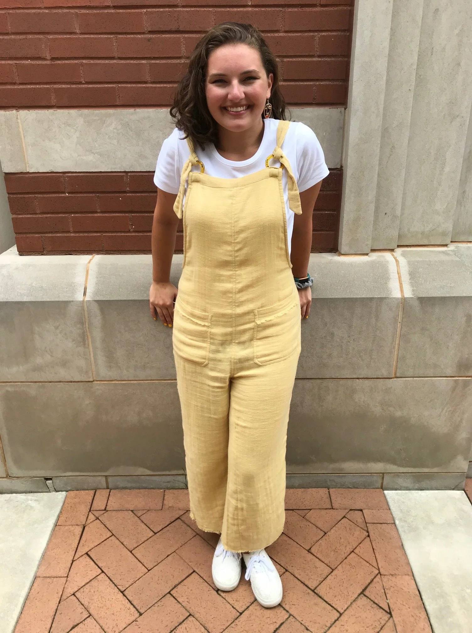 A young woman with wavy brown hair smiling and standing outdoors in front of a brick and concrete wall, wearing a yellow overall skirt over a white t-shirt and white sneakers.