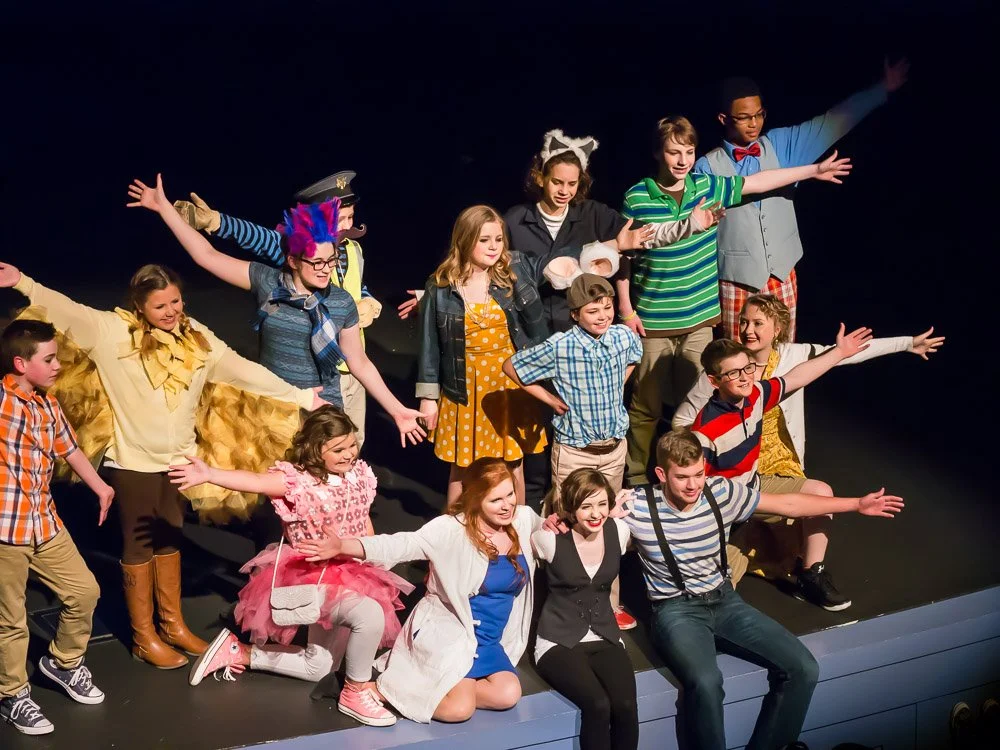 A group of children performing on stage, dressed in colorful costumes, with some wearing animal ears and others in vibrant clothing, striking various poses with arms outstretched.