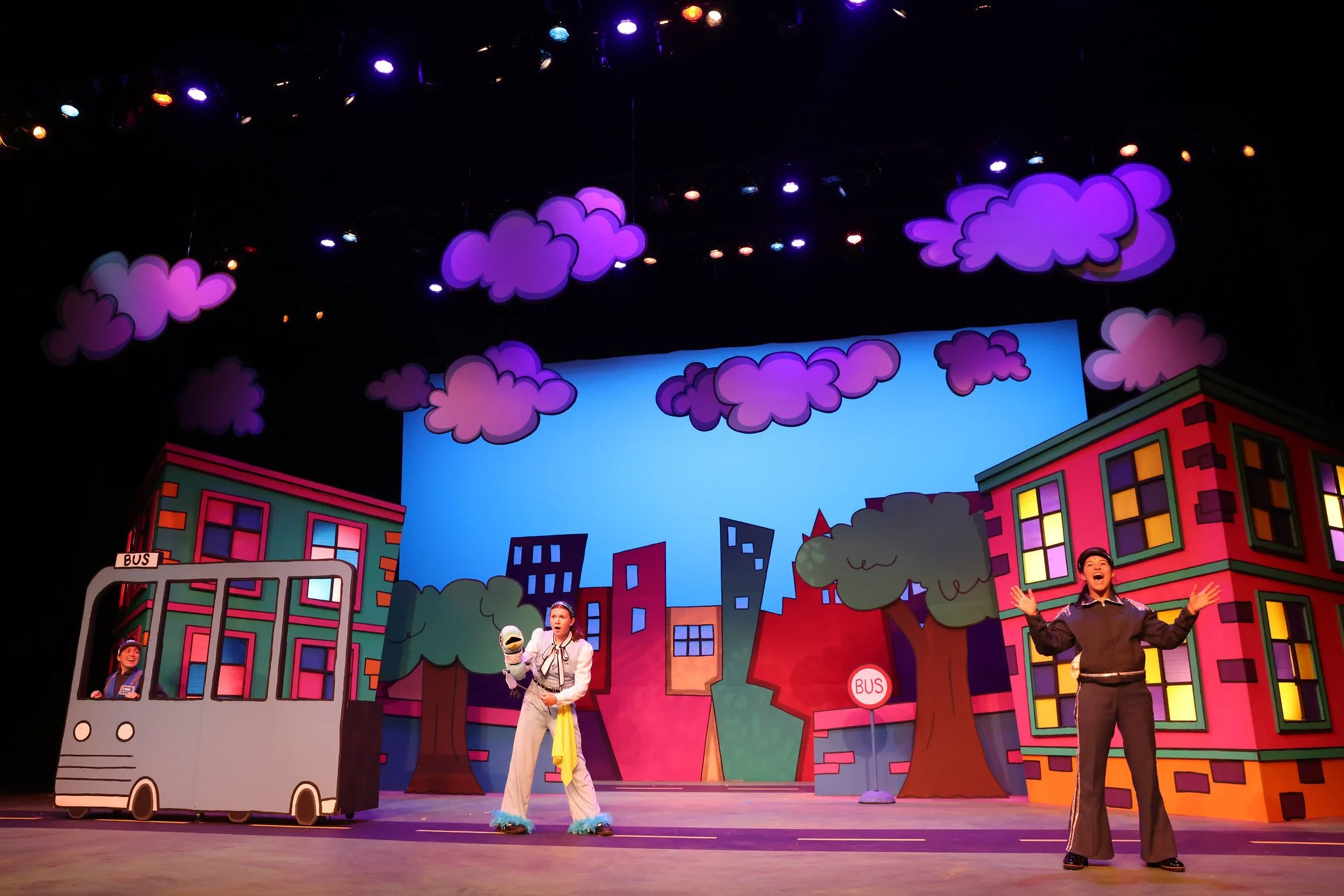 Stage scene from a children's theatrical production with colorful animated cityscape backdrop, purple clouds hanging overhead, and performers in costumes including a puppet and an adult actor with open arms.