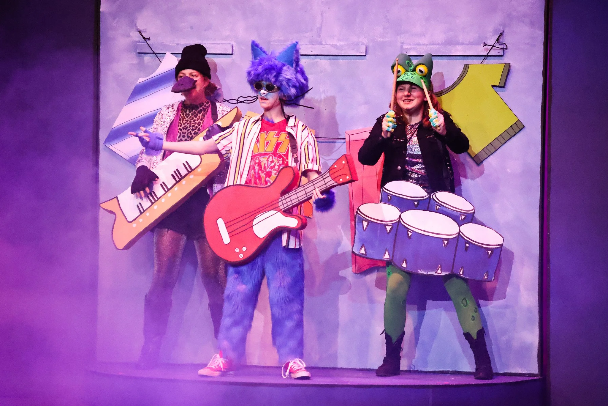 Three people dressed in colorful, playful costumes stand on a stage. The person on the left wears a black beanie, a long-sleeve top with leopard print, and holds a cardboard guitar. The person in the middle is in a wolf costume with blue fur, sunglasses, and a striped shirt. The person on the right wears a green lizard mask, gloves, and holds drumsticks, standing behind painted cardboard drums. The background has a purple hue with large, cartoonish block letters.