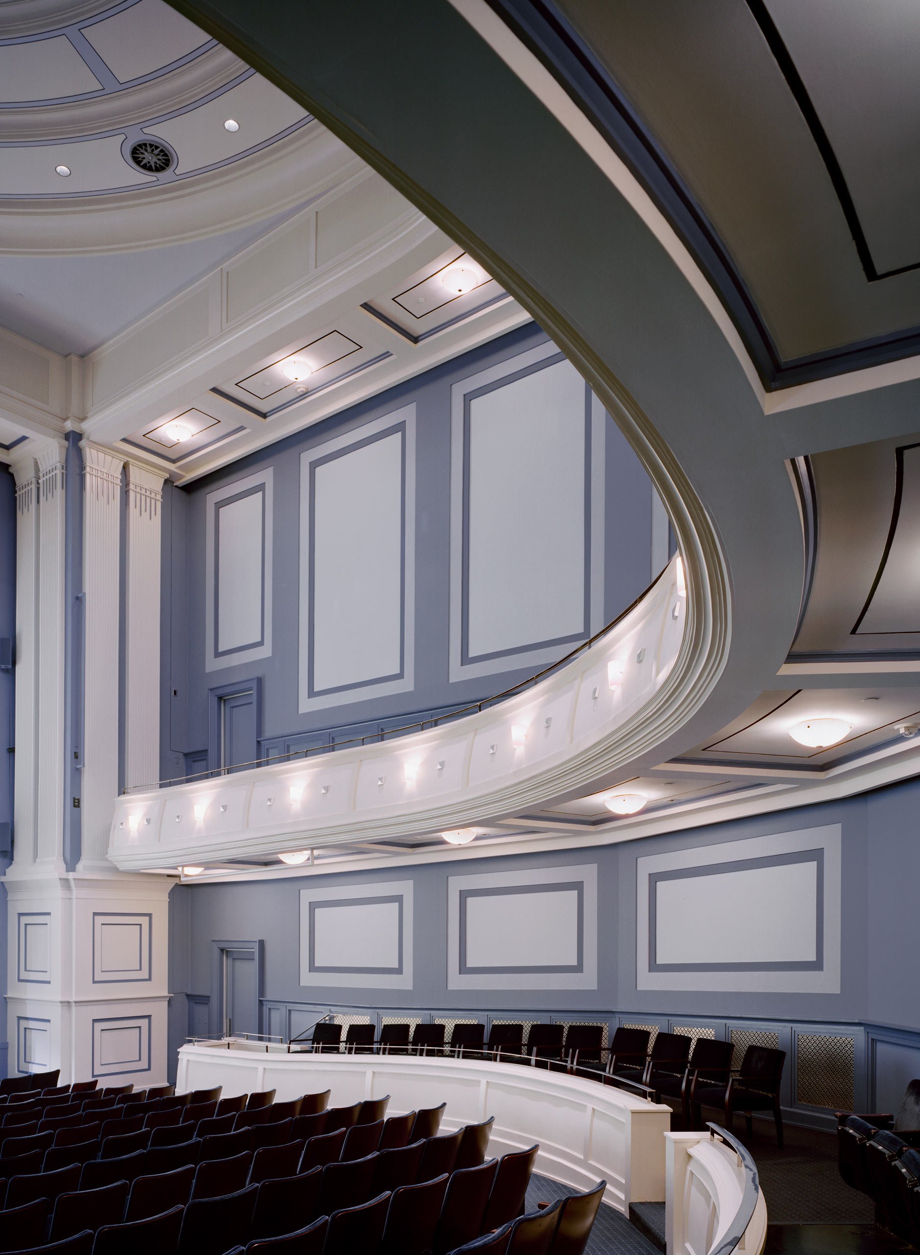 Interior view of a theater auditorium with blue walls, white trim, and black seats arranged in multiple rows. There is a balcony with additional seating and decorative molding along the ceiling.