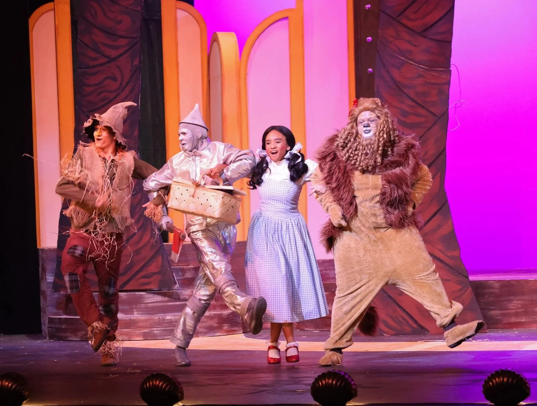 Actors in colorful costumes performing a scene on stage with a pink and purple lit background, including a lion, a scarecrow, and children.