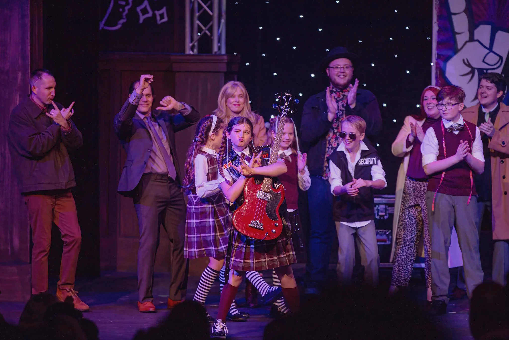 A group of children and adults on stage at a theatrical or musical performance. The children are dressed in colorful costumes, with one girl holding a red electric guitar and making a peace sign. The adults are clapping and smiling. The background has starry lights and show banners.