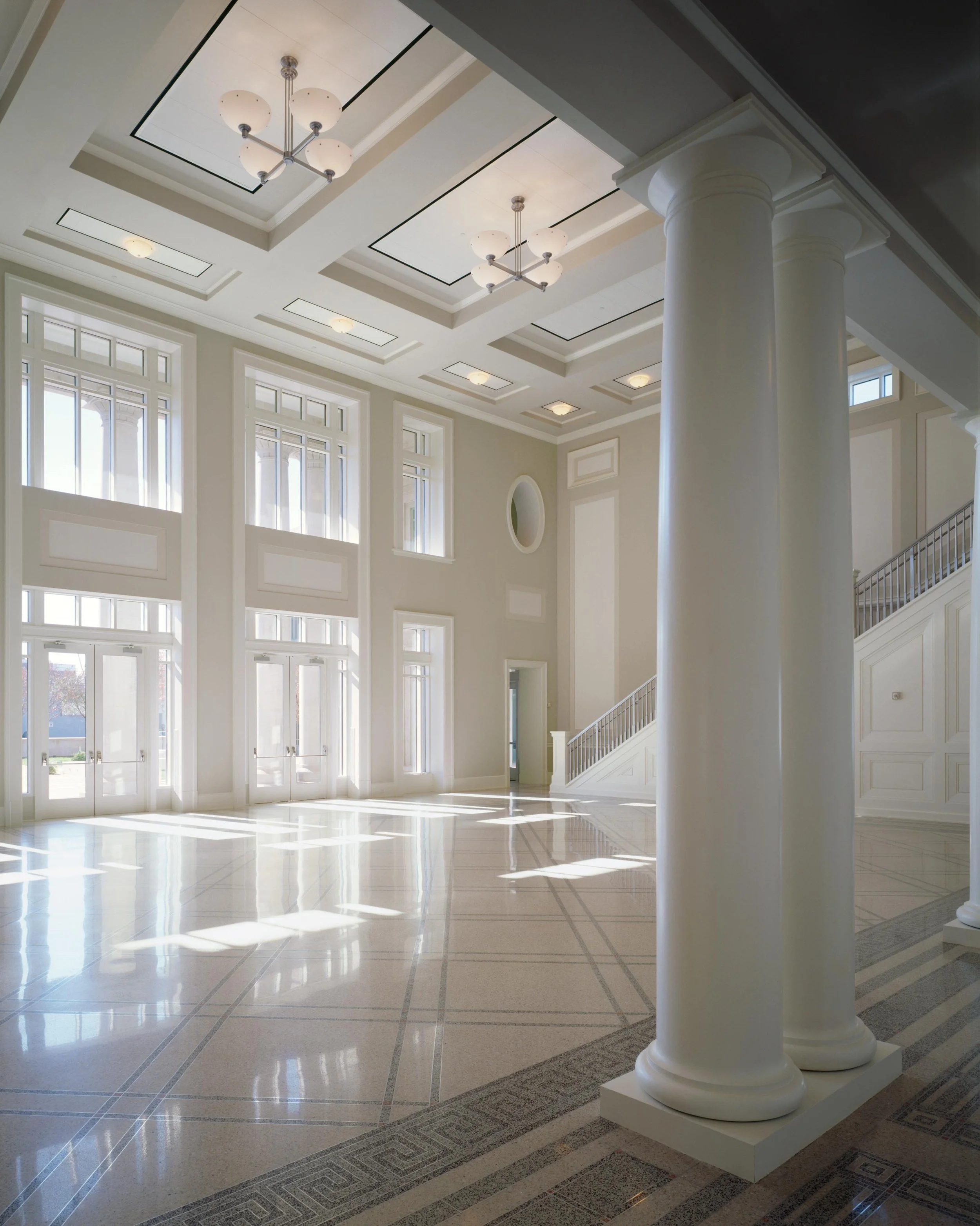 A spacious, well-lit interior with large windows, high ceiling, and elegant white columns, reflecting sunlight on the polished tiled floor.