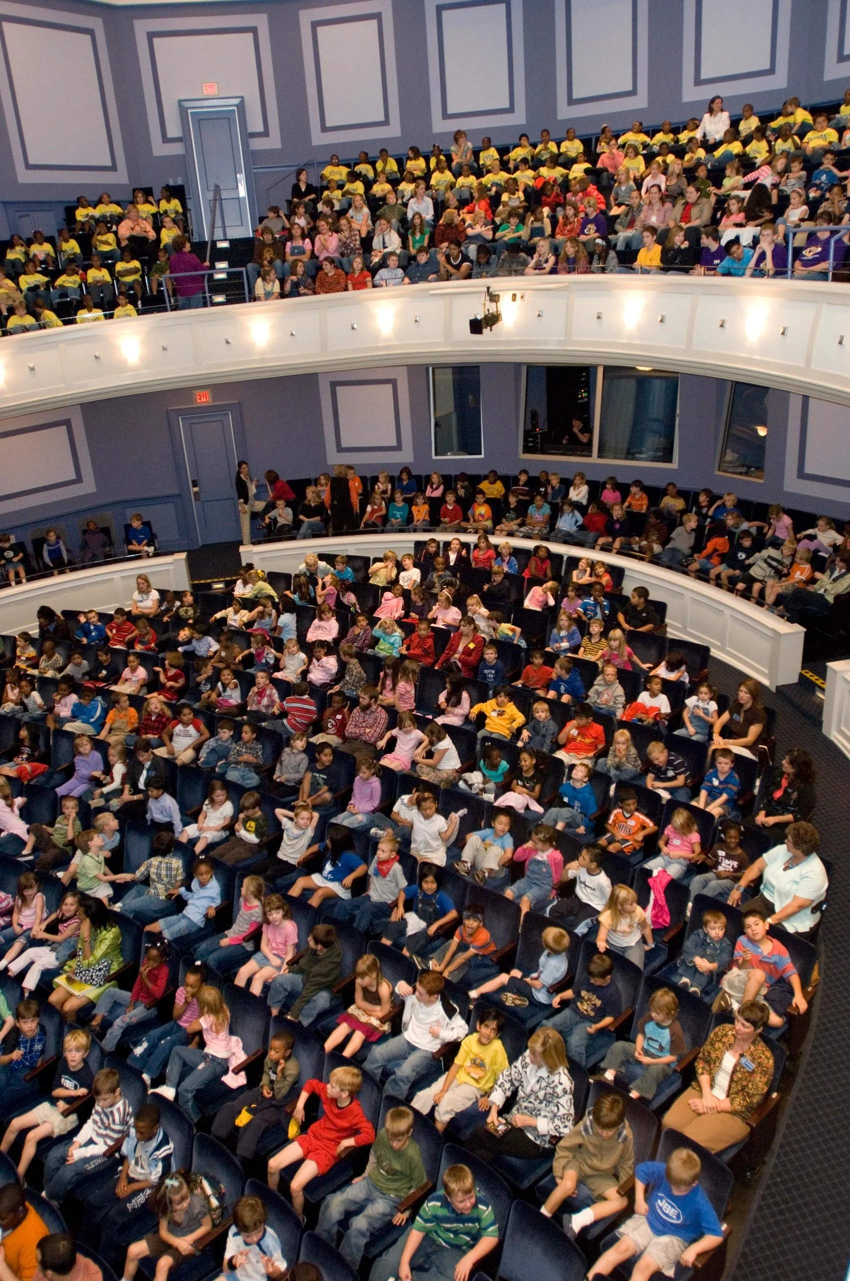 Large auditorium filled with children and some adults seated in curved rows of chairs, some wearing yellow shirts, others in various casual clothing, with a blue wall and windows at the back.