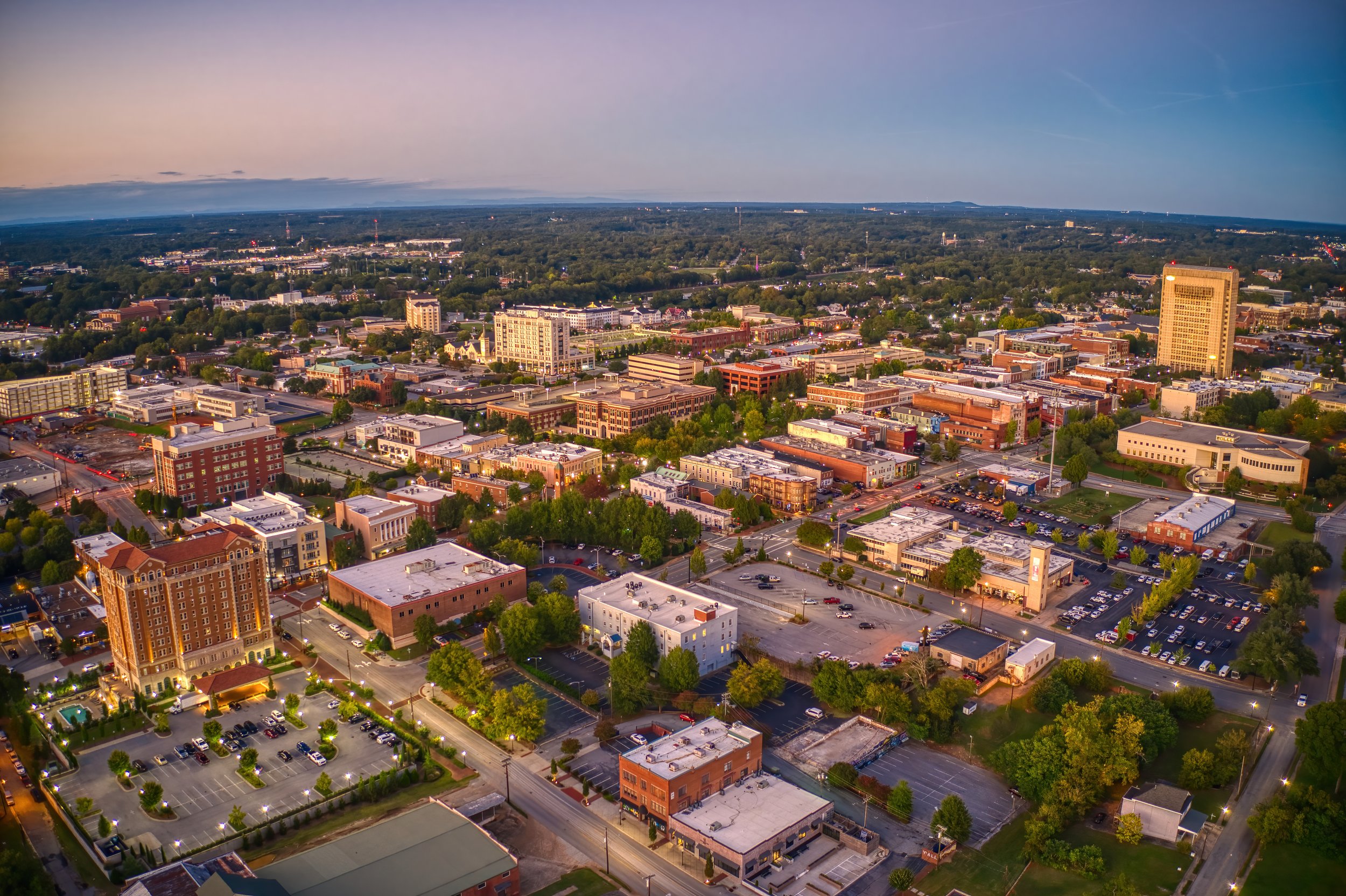 Aerial view of a cityscape at dusk with a mix of tall offices, smaller buildings, parking lots, trees, and streets stretching into the horizon under a pinkish sky.
