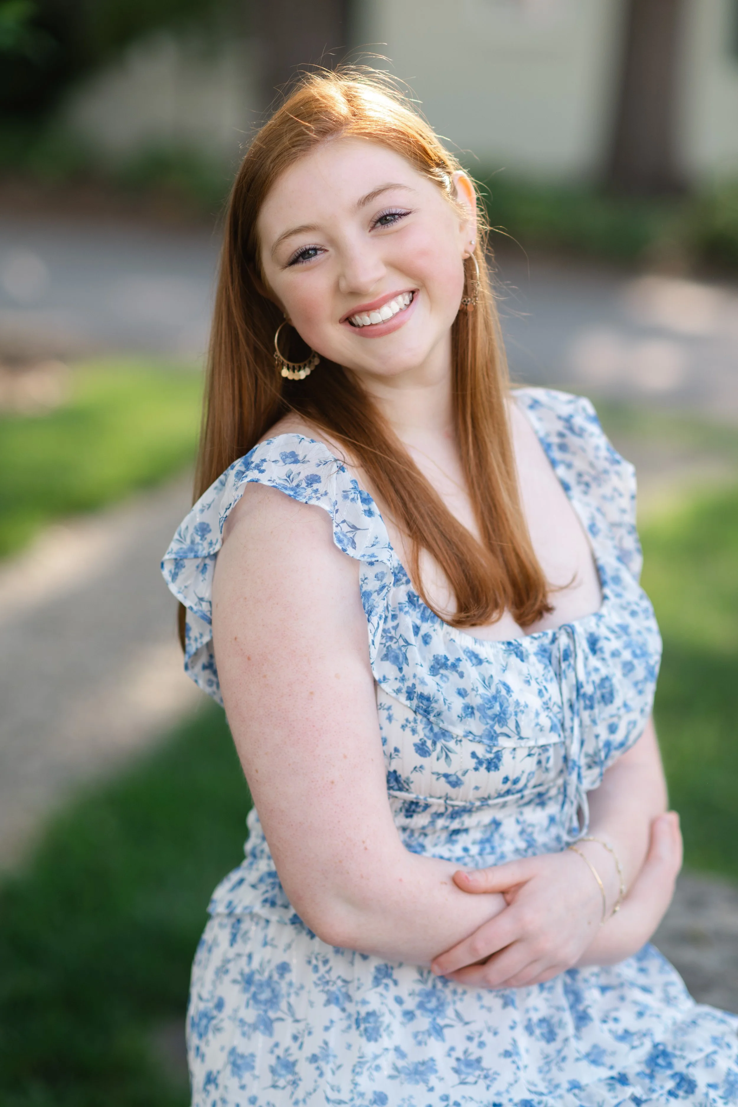 A red-haired woman smiling outdoors in sunlight, wearing a blue and white floral dress with ruffled sleeves and hoop earrings.