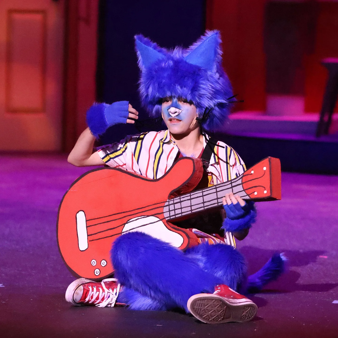 Person in a blue furry animal costume with large ears, face paint, and red sneakers, sitting on the stage with a large red cardboard guitar, during a performance.