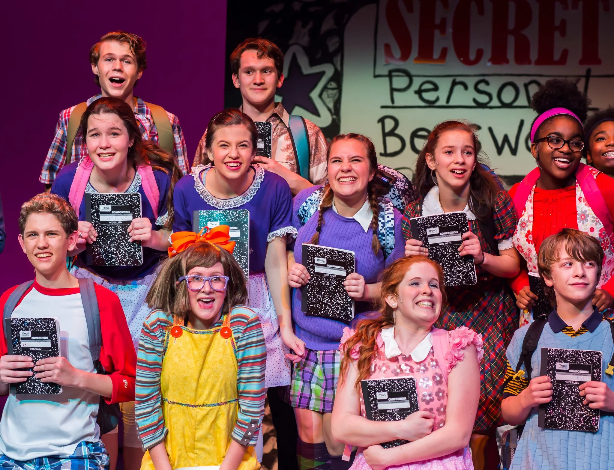 Group of children standing on stage, smiling, holding notebooks, with a colorful background and a sign that says 'Teacher Person'.
