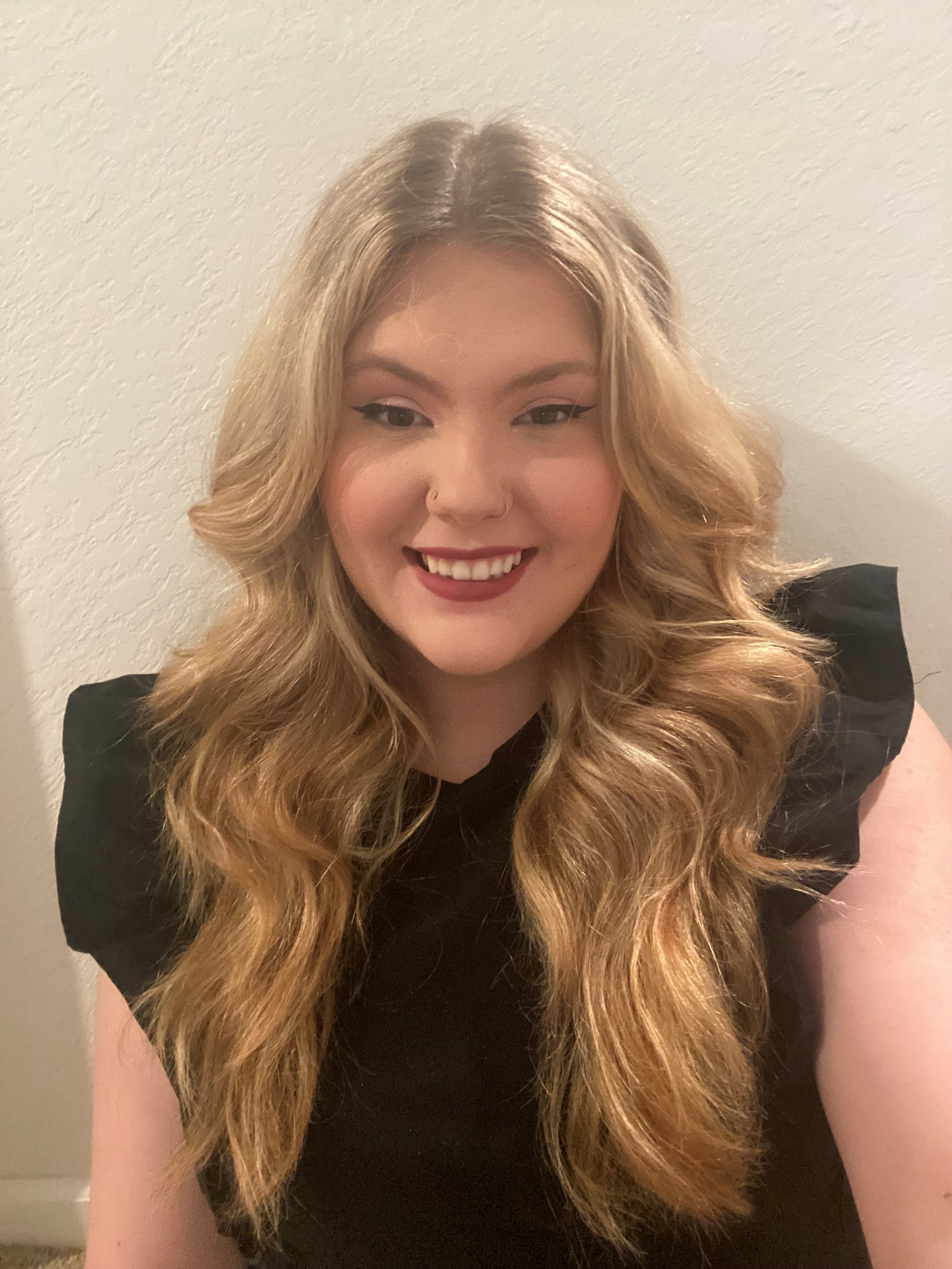 A woman with long, wavy blonde hair smiling and wearing a black top, standing against a plain light-colored wall.