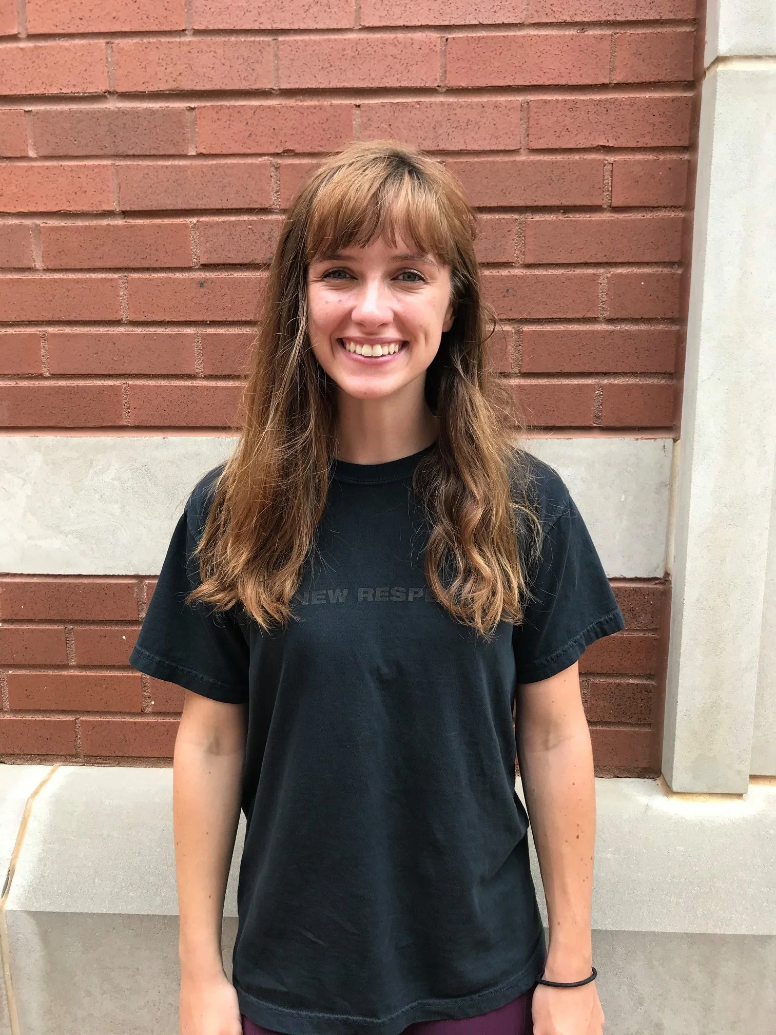 Young woman with long brown hair and a big smile, wearing a black t-shirt, standing outdoors in front of a red brick wall.