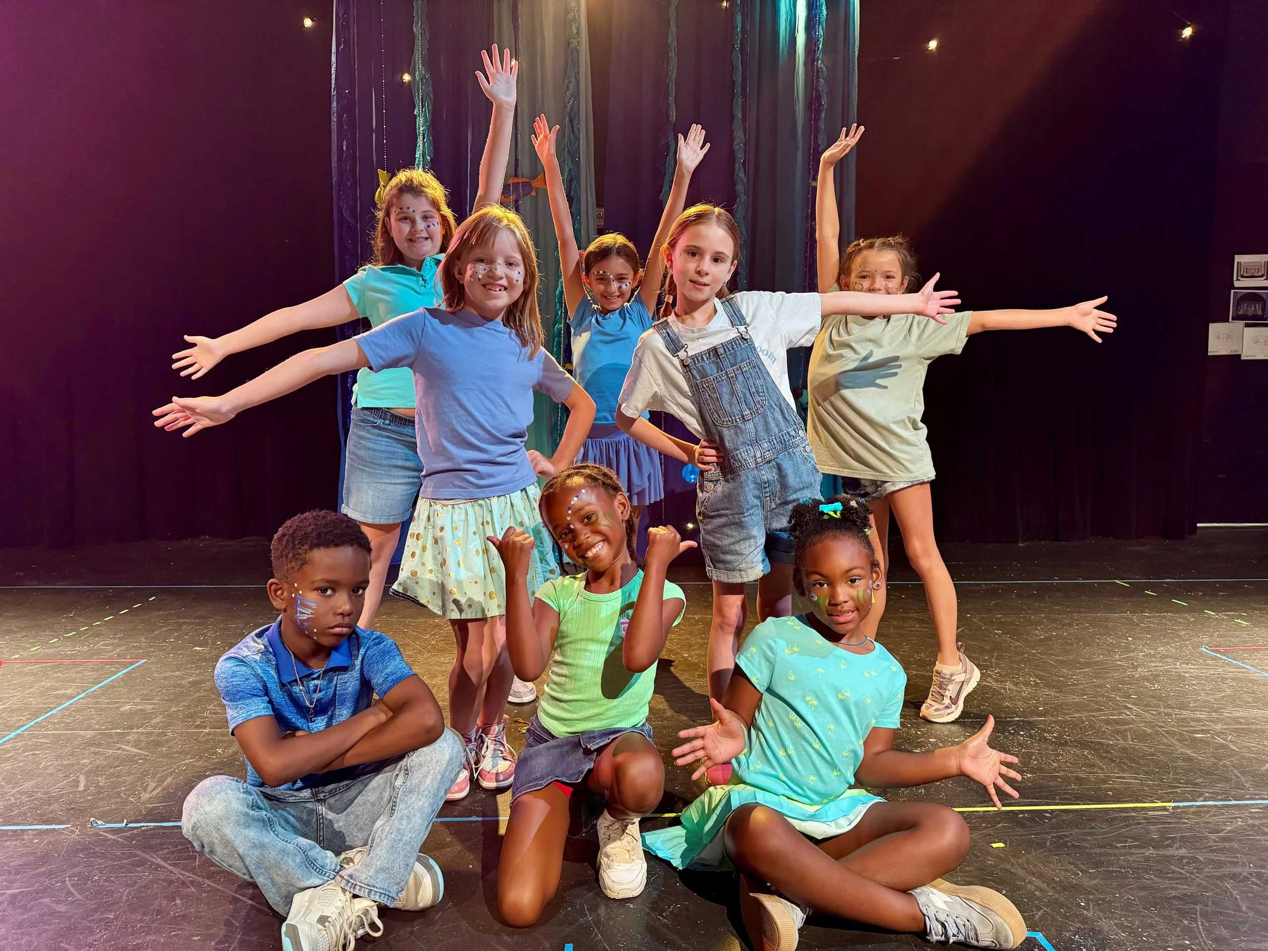 Group of six girls and three boys on stage, some with painted faces, some with arms raised or outstretched, in front of dark curtains with string lights, posing for a photo.