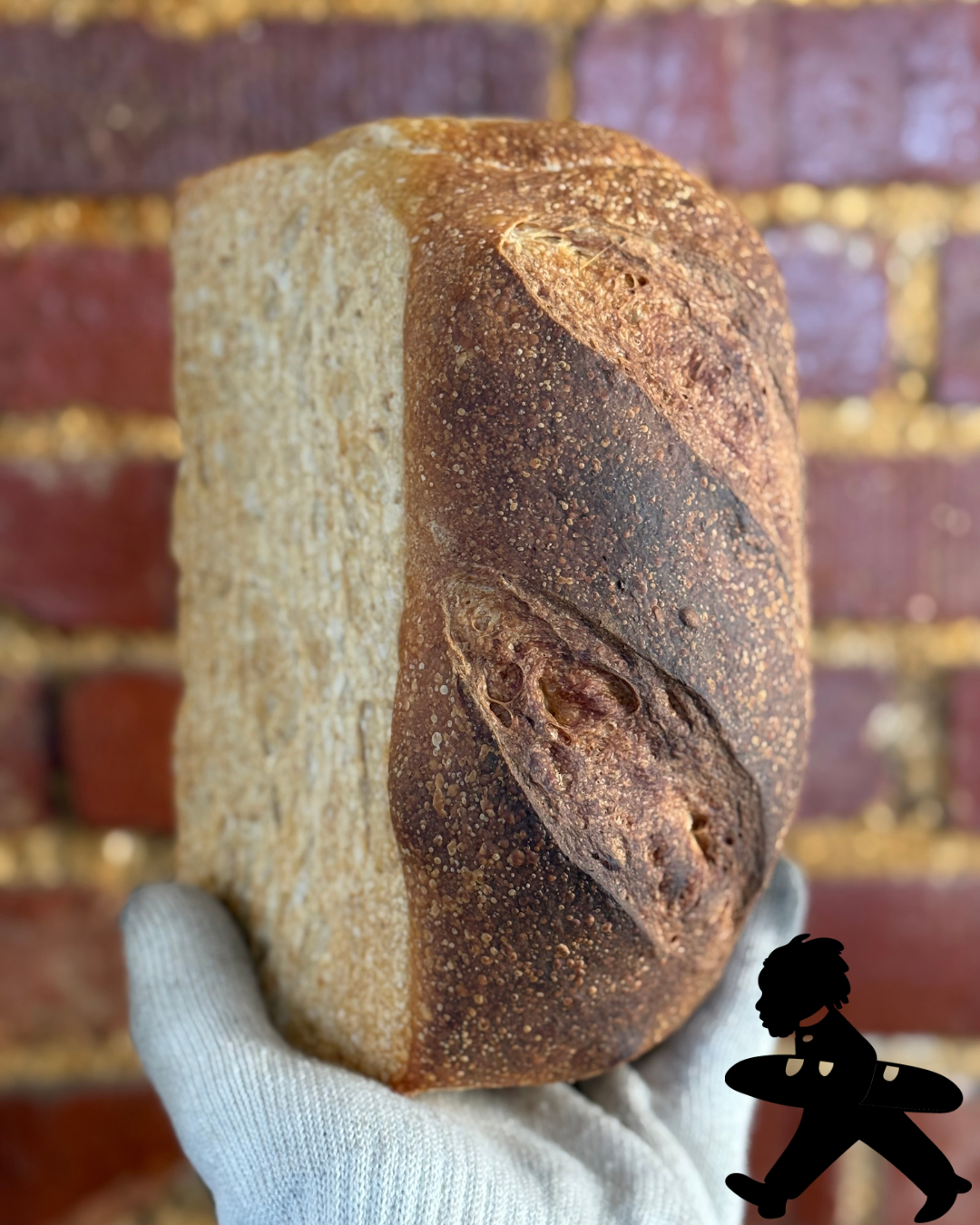 A person holding a piece of rustic, crusty bread with a darker crust on one side and a lighter, softer interior on the other, against a red brick wall background.