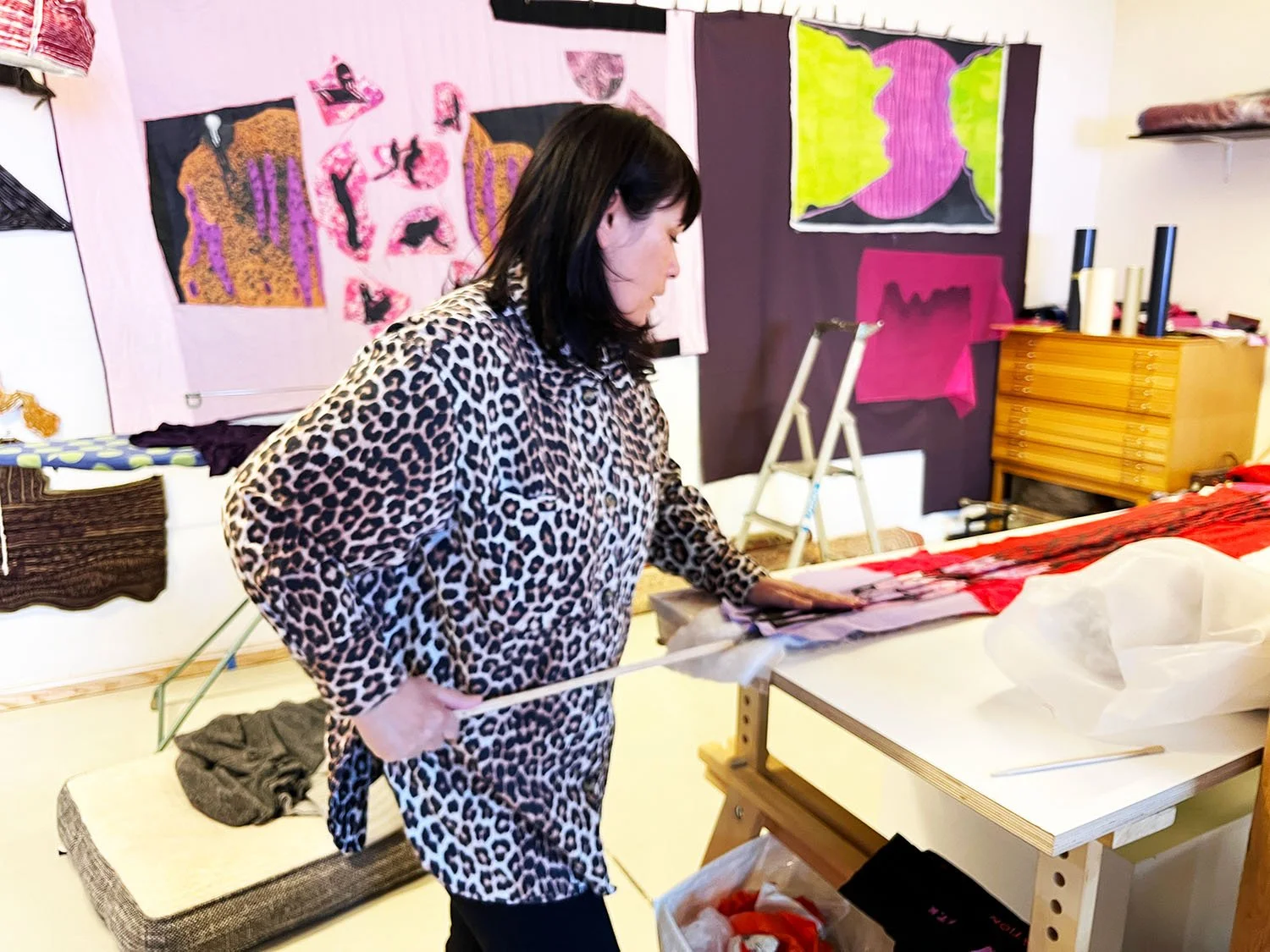 A woman in a leopard print dress is standing at a table with fabric, in an art studio with colorful artwork on the walls.