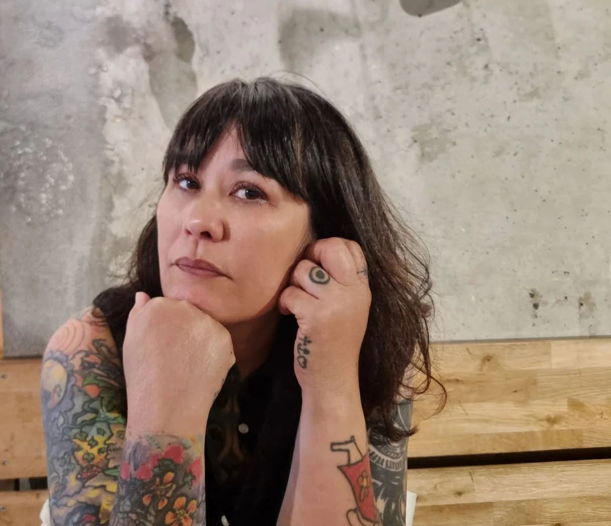 A woman with dark hair and tattoos rests her chin on her hand, looking at the camera, in front of a wooden surface and concrete wall.