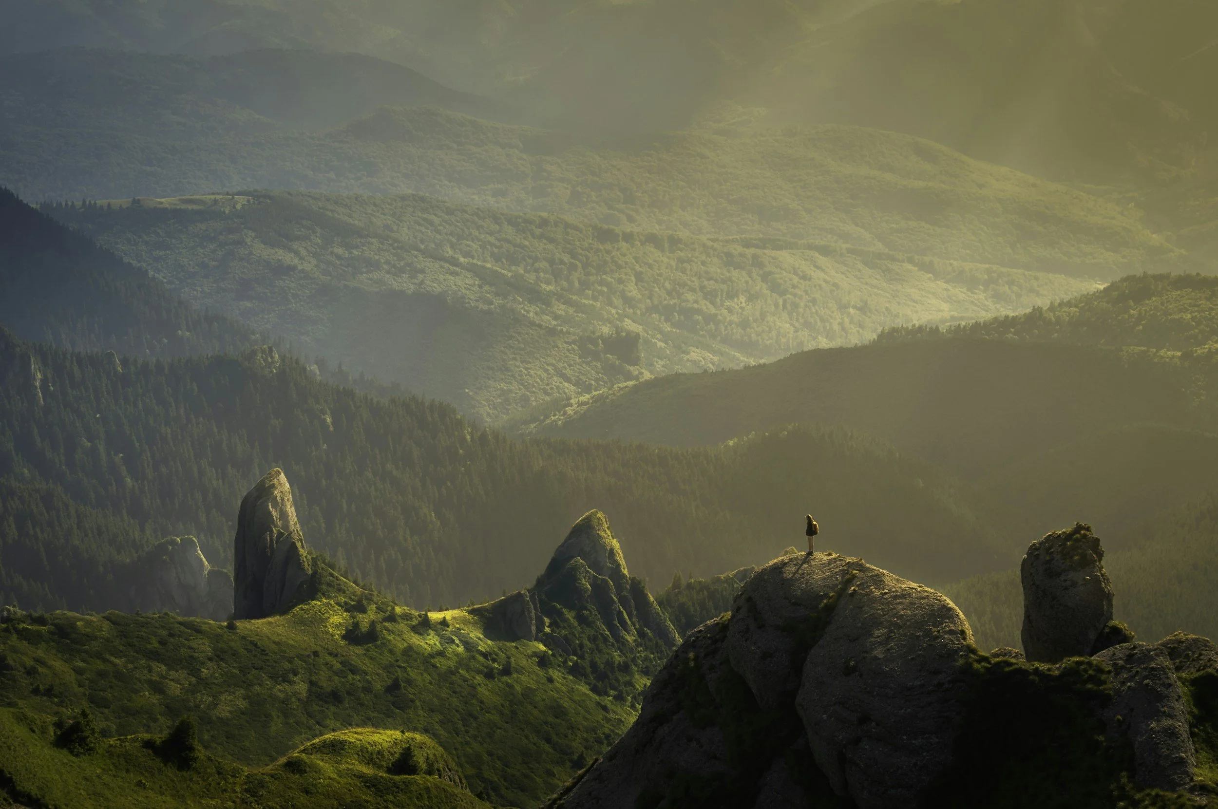 A person standing on a rocky ledge overlooking a lush green valley with rolling hills and mountain ranges in the distance, lit by warm sunlight.