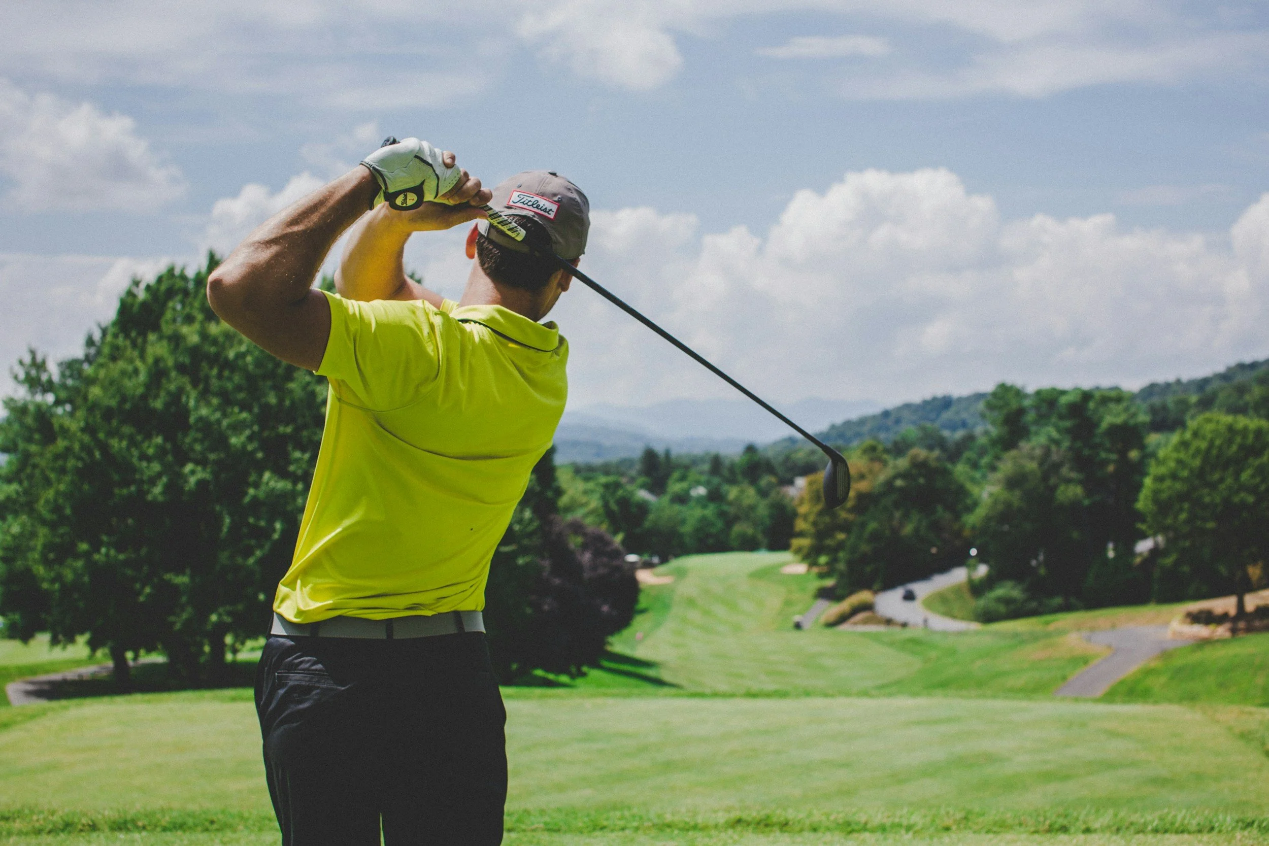 A man in a yellow shirt and gray cap swings a golf club on a golf course with lush green grass and trees, under a partly cloudy sky.