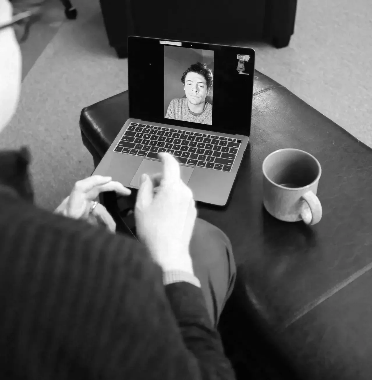 A person sitting at a table holding a smartphone, during a video call on a laptop showing a man with curly hair in a gray shirt. A coffee mug is on the table.