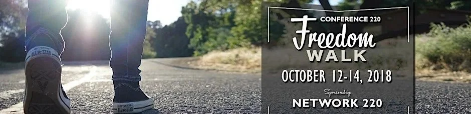 Close-up of a person's feet walking outdoors on a paved trail during daytime with sunlight, accompanied by a promotional graphic for 'Conference 220 Freedom Walk' held October 12-14, 2018, sponsored by Network 220.