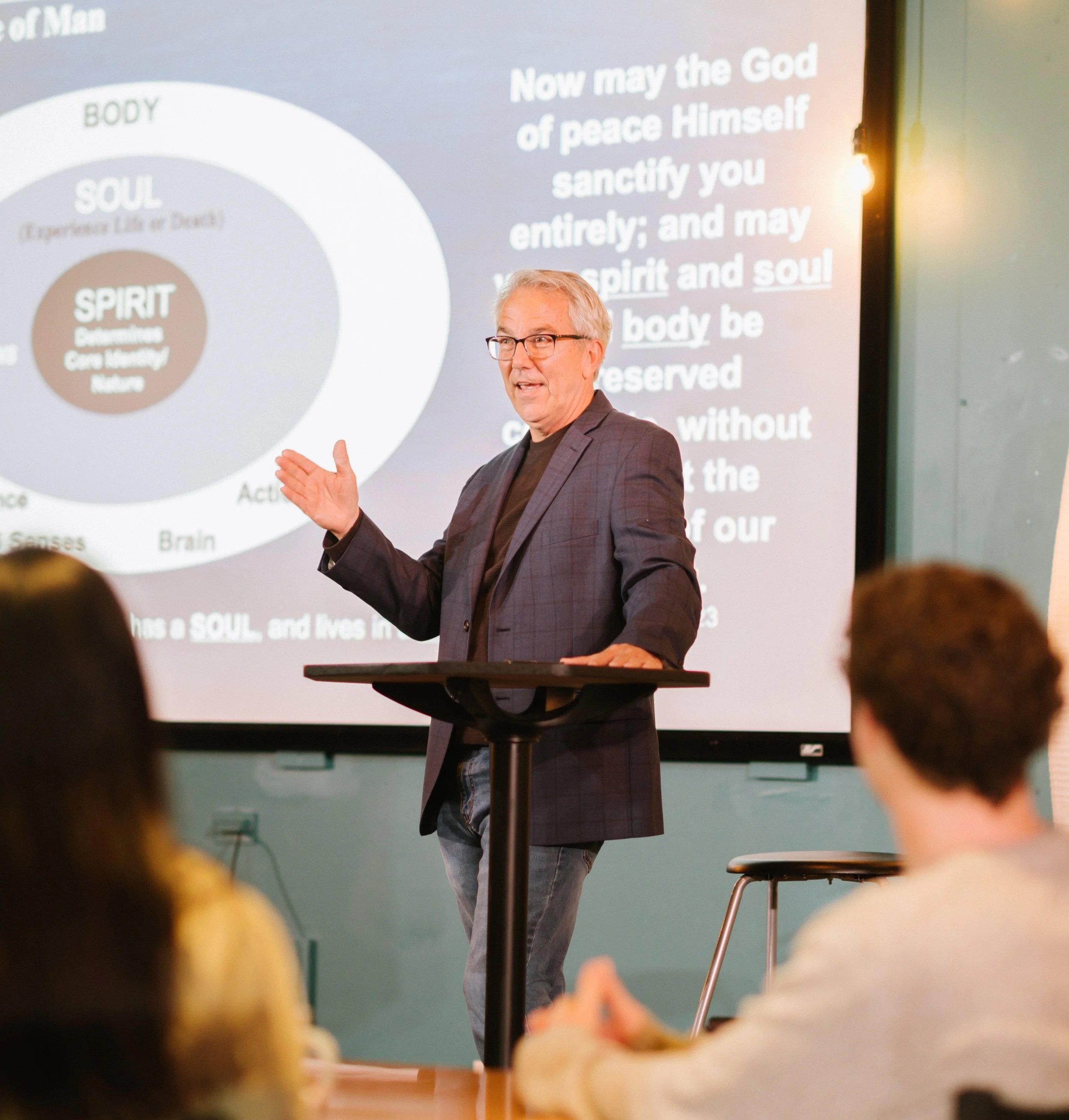 A man in a grey blazer and glasses giving a presentation in front of a large screen with a diagram and text, while an audience listens.