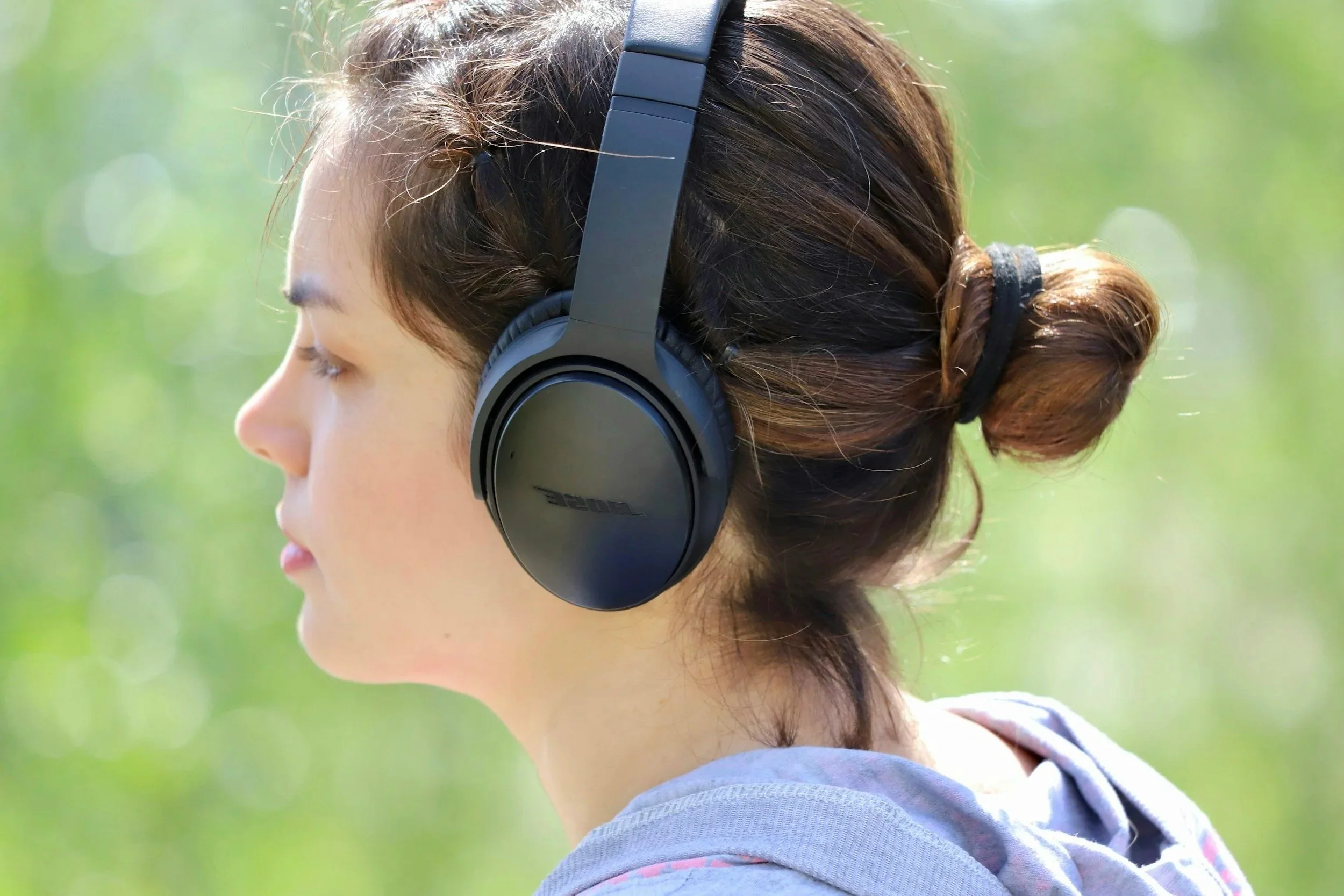 A young woman is outdoors wearing black Bose headphones with her eyes closed and hair tied back in a bun, with a blurred green background.