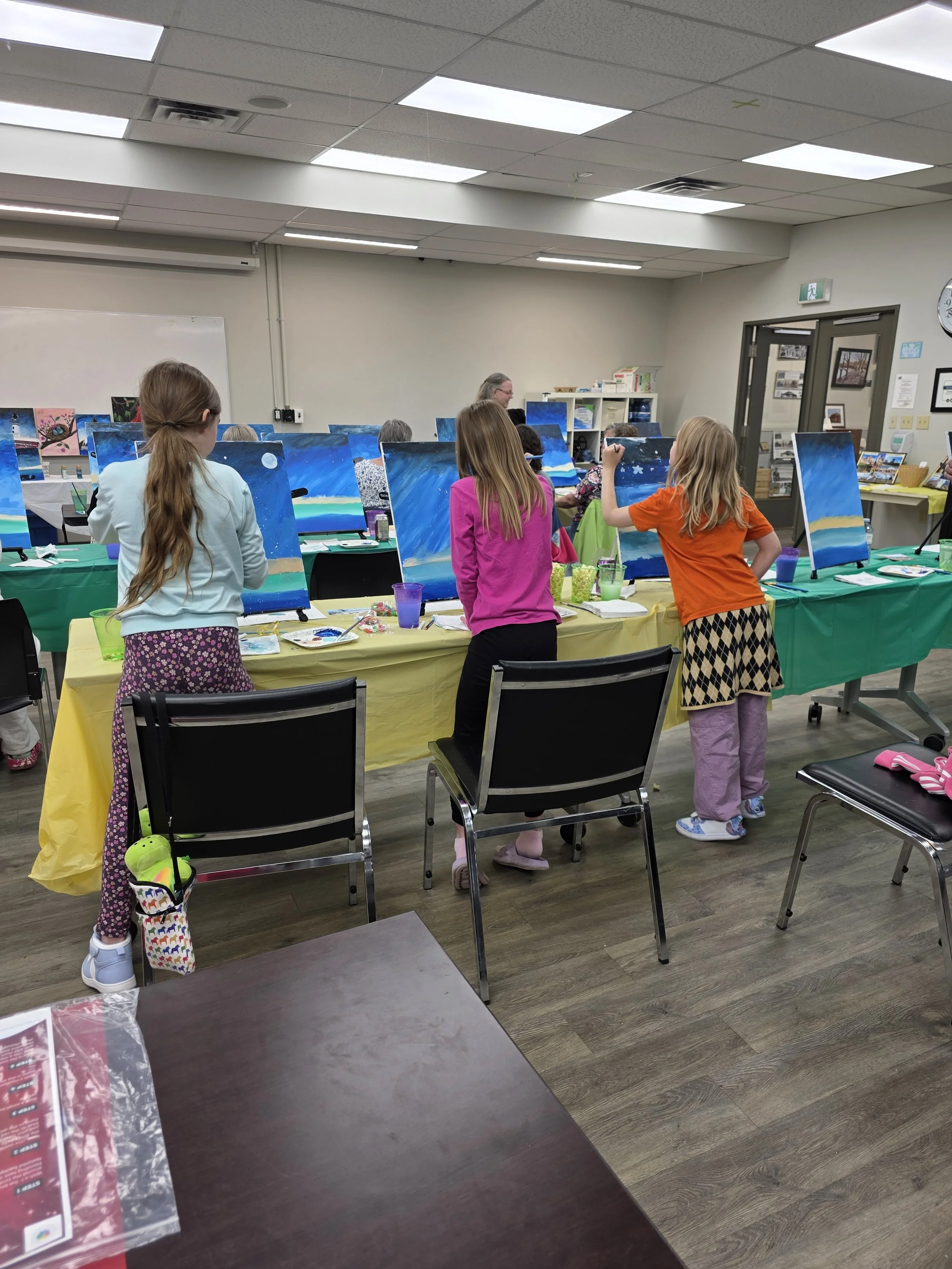 Children and adults painting landscapes on canvases at a group art class in a community room.
