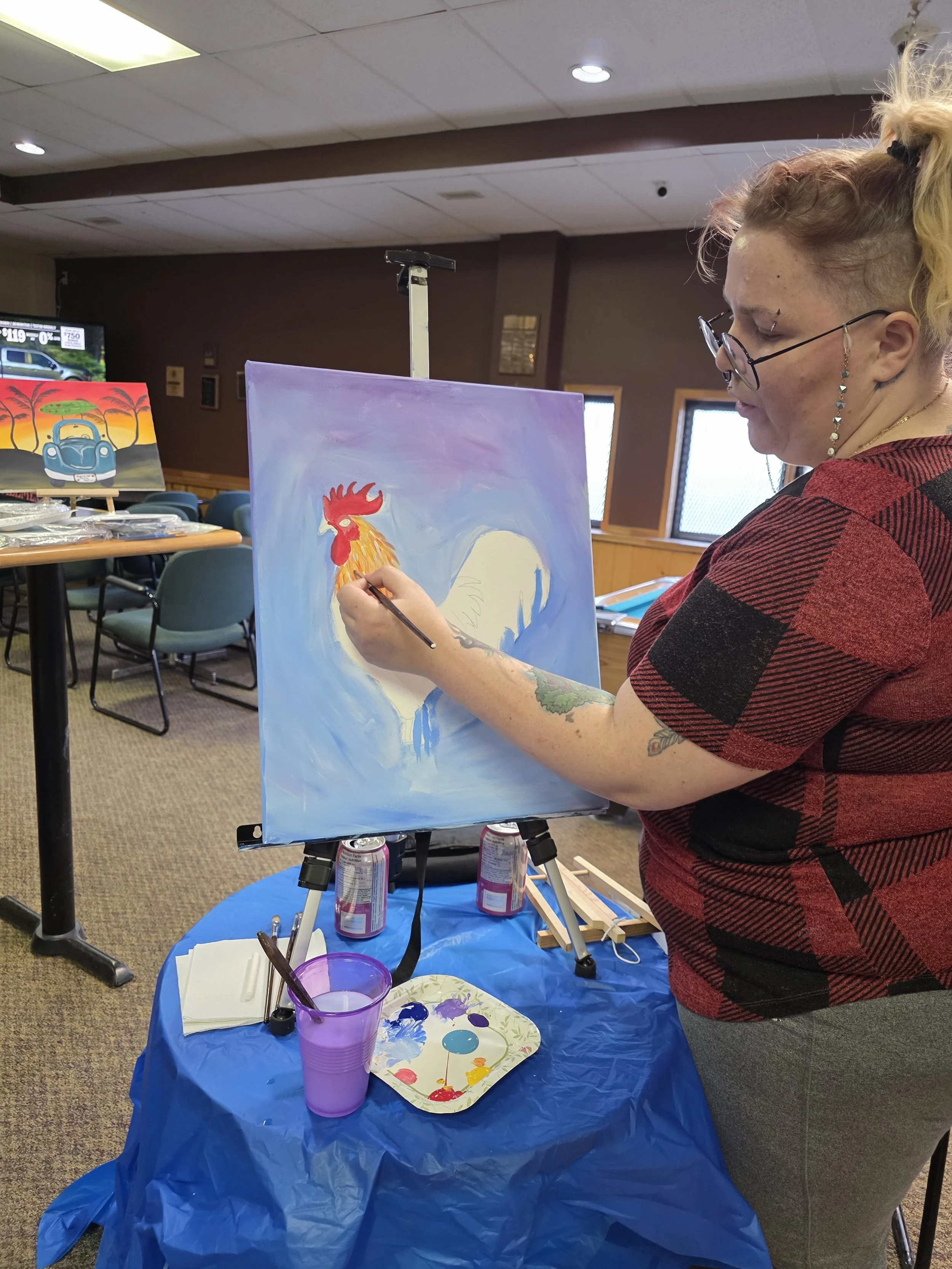 A woman with blonde hair tied back, wearing glasses, painted a rooster on a canvas at an art event. The canvas is on an easel, and art supplies are on a table covered with a blue plastic sheet, including paints, brushes, and cans of soda. In the background, there are chairs and a colorful painting of a car with a sunset sky.