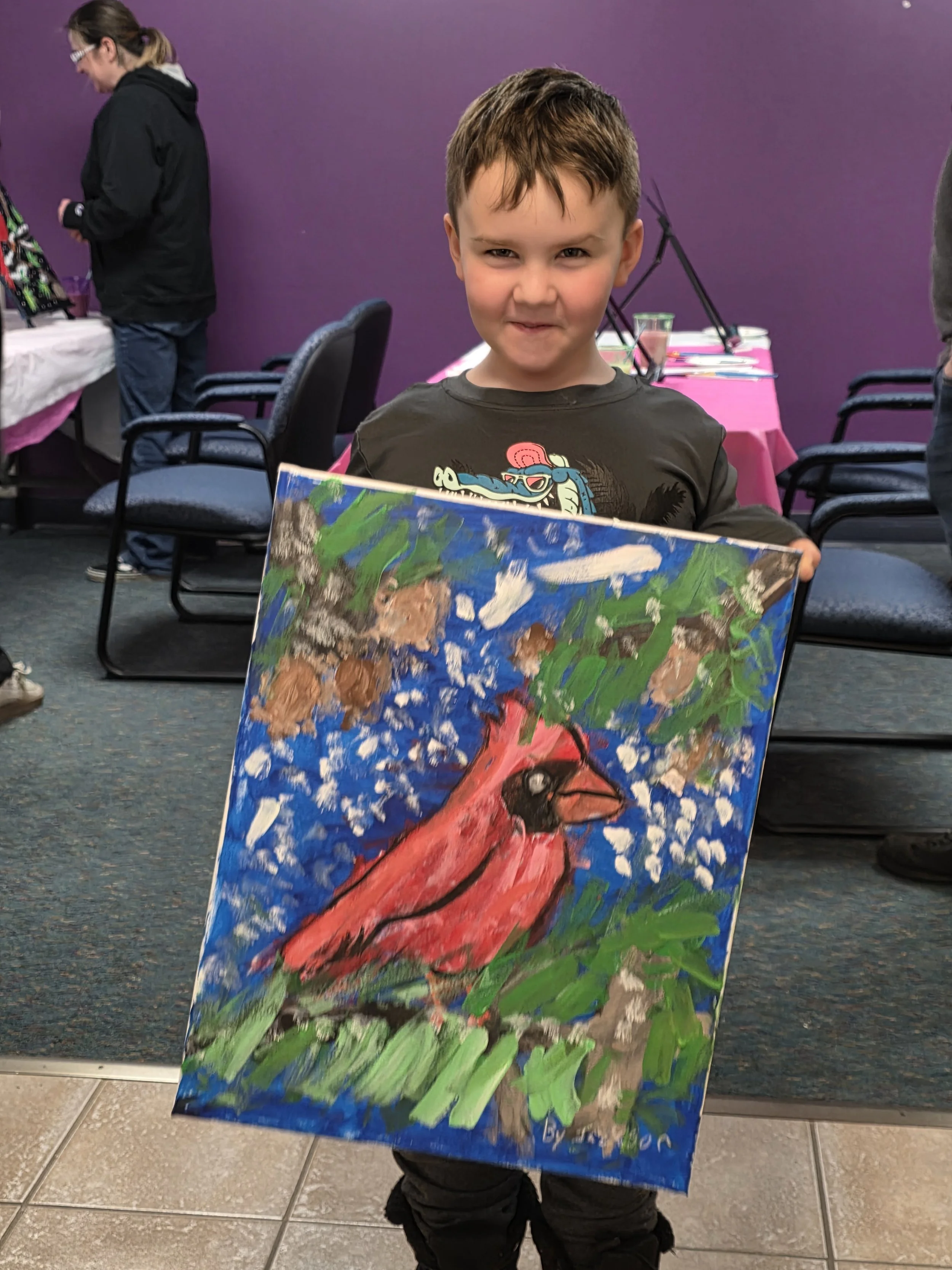 A young boy holding a colorful painting of a pink flamingo standing on green grass. He is standing indoors with a purple wall behind him and a table with craft supplies in the background.