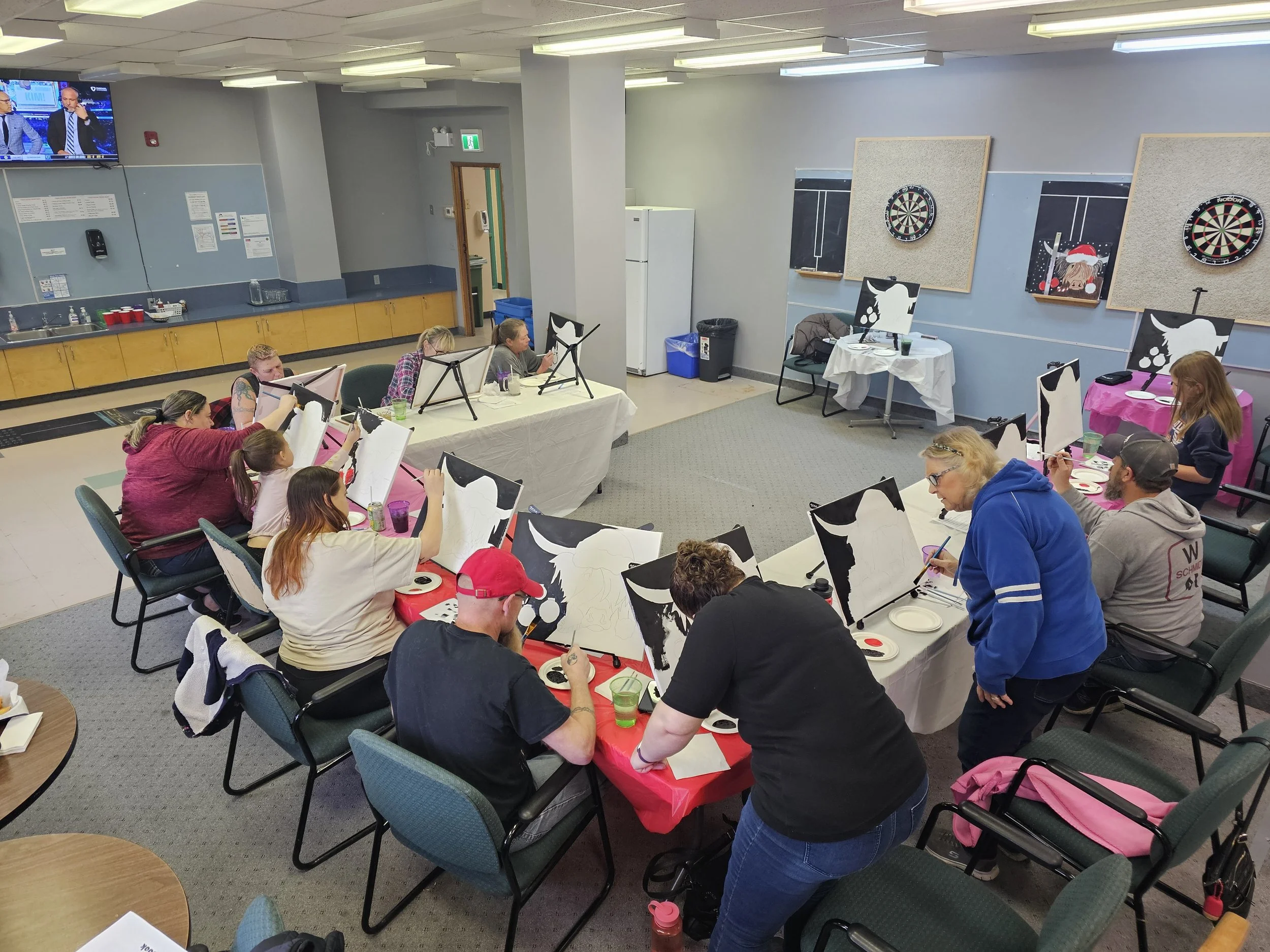 People participating in a painting event, sitting at tables with painting canvases and supplies, in a community room decorated with dartboards and holiday-themed paintings.