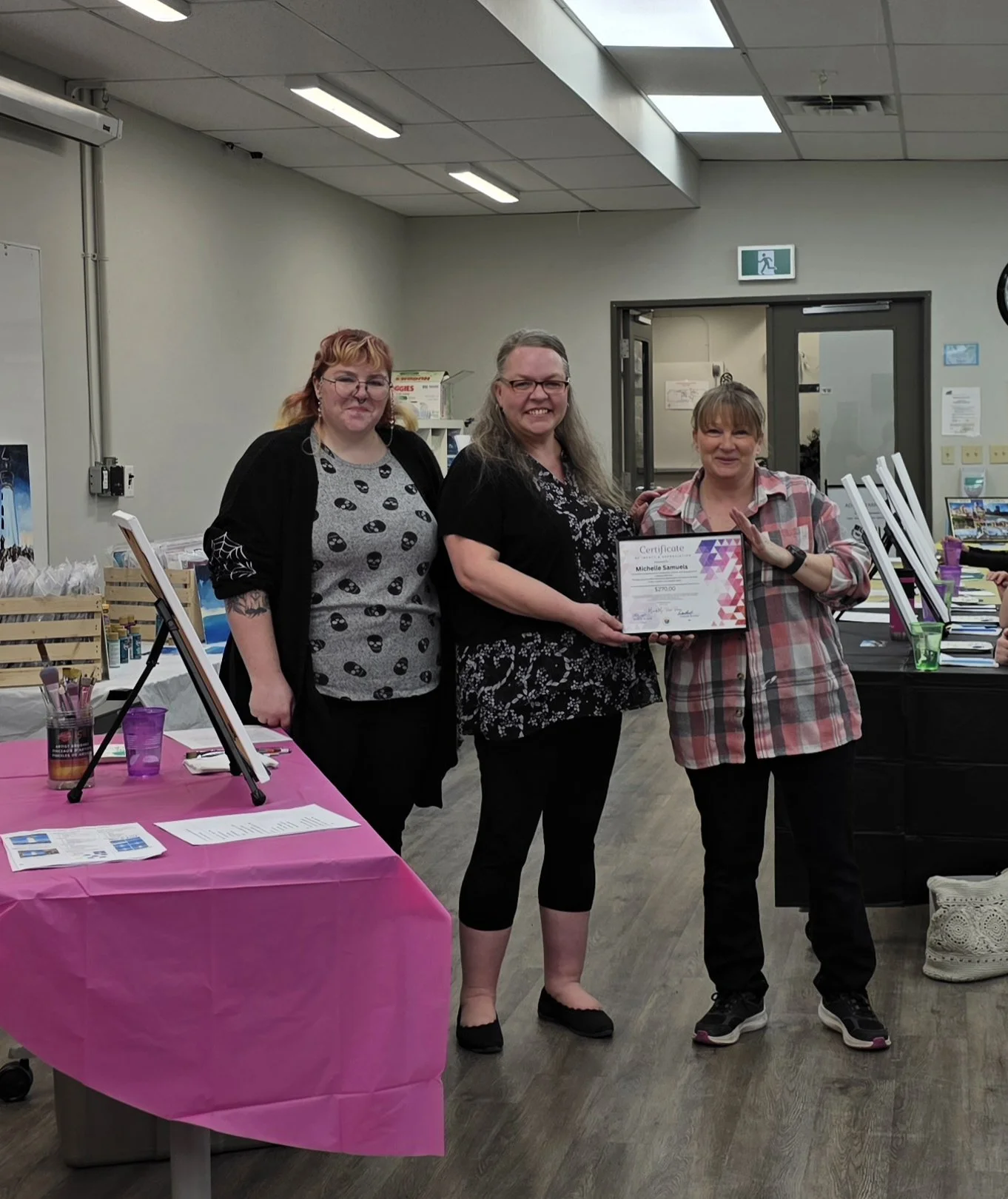 Three women standing together at an indoor event, with one woman holding a certificate. A table with pink tablecloth and display items is visible on the left side of the image. The room has fluorescent lighting and a door in the background.