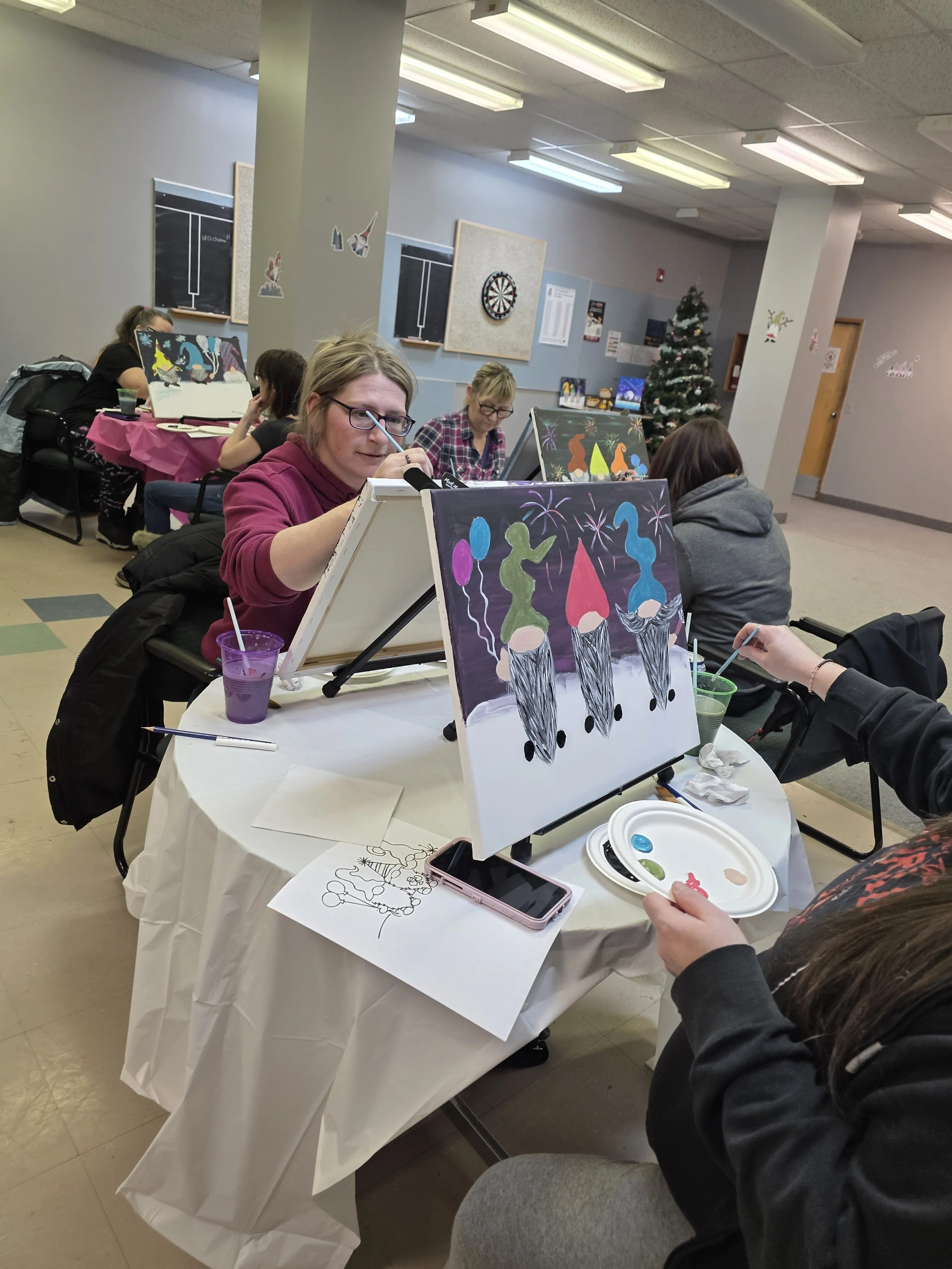 People sitting at a table painting holiday-themed pictures, with a Christmas tree and holiday decorations in the background.