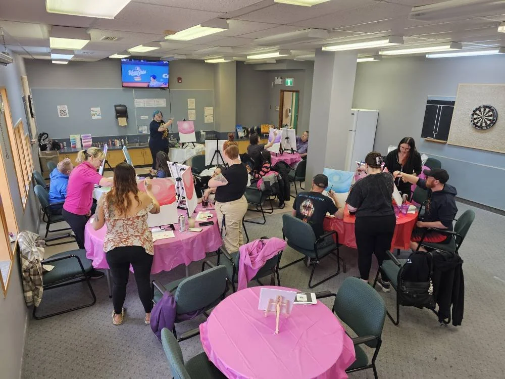A room filled with women painting on canvas boards at pink-covered tables during a painting class or workshop.