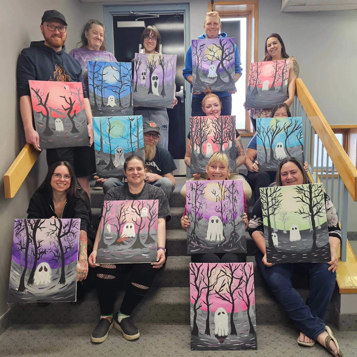 Group of people on stairs holding colorful paintings of spooky halloween-themed trees and ghostly figures in various shades of purple, pink, blue, and gray.
