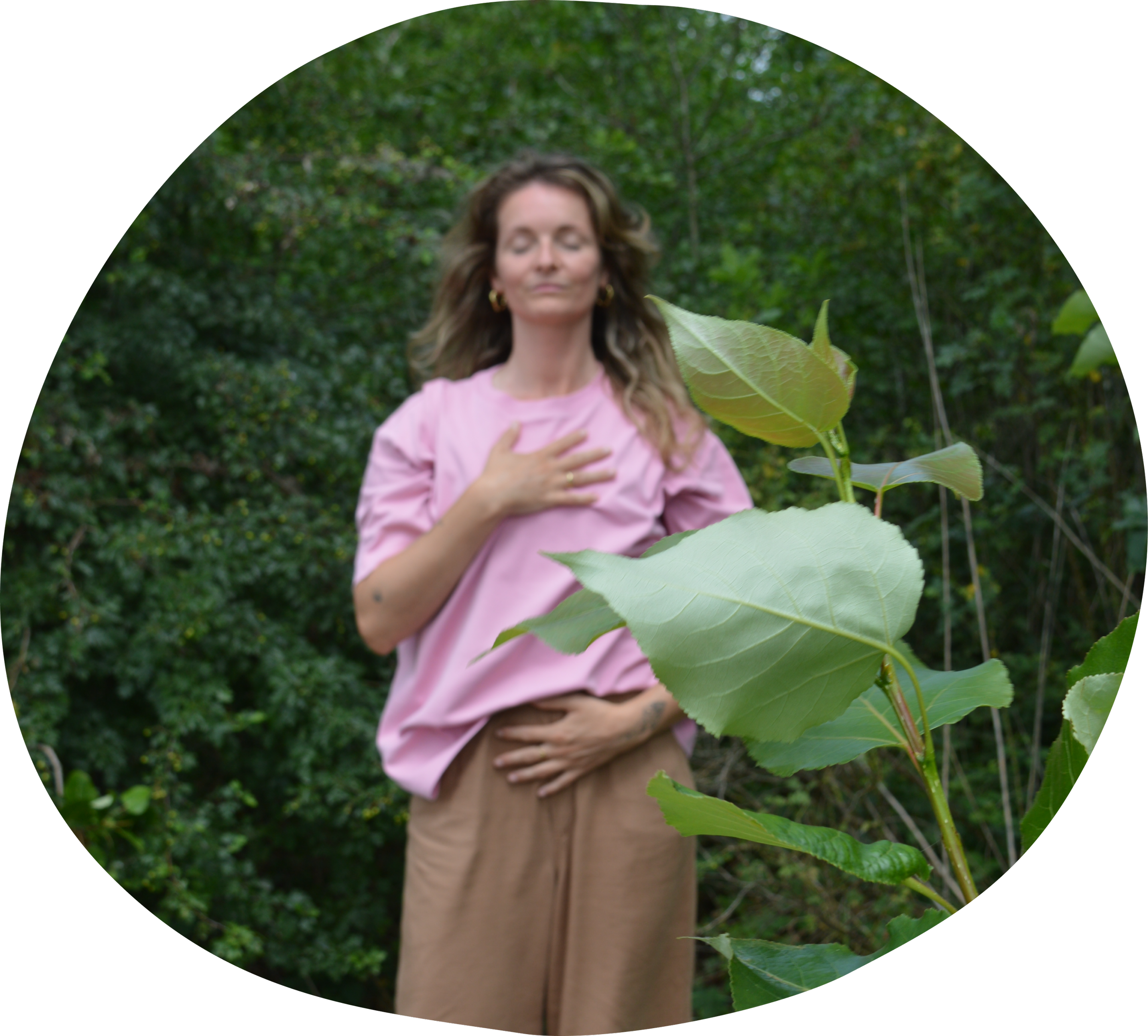 Nadia van Wylde Roots met lang haar in een bos, met een roze trui en een beige broek, zacht ademend, hand op haar borst, grote groene bladeren voor haar.